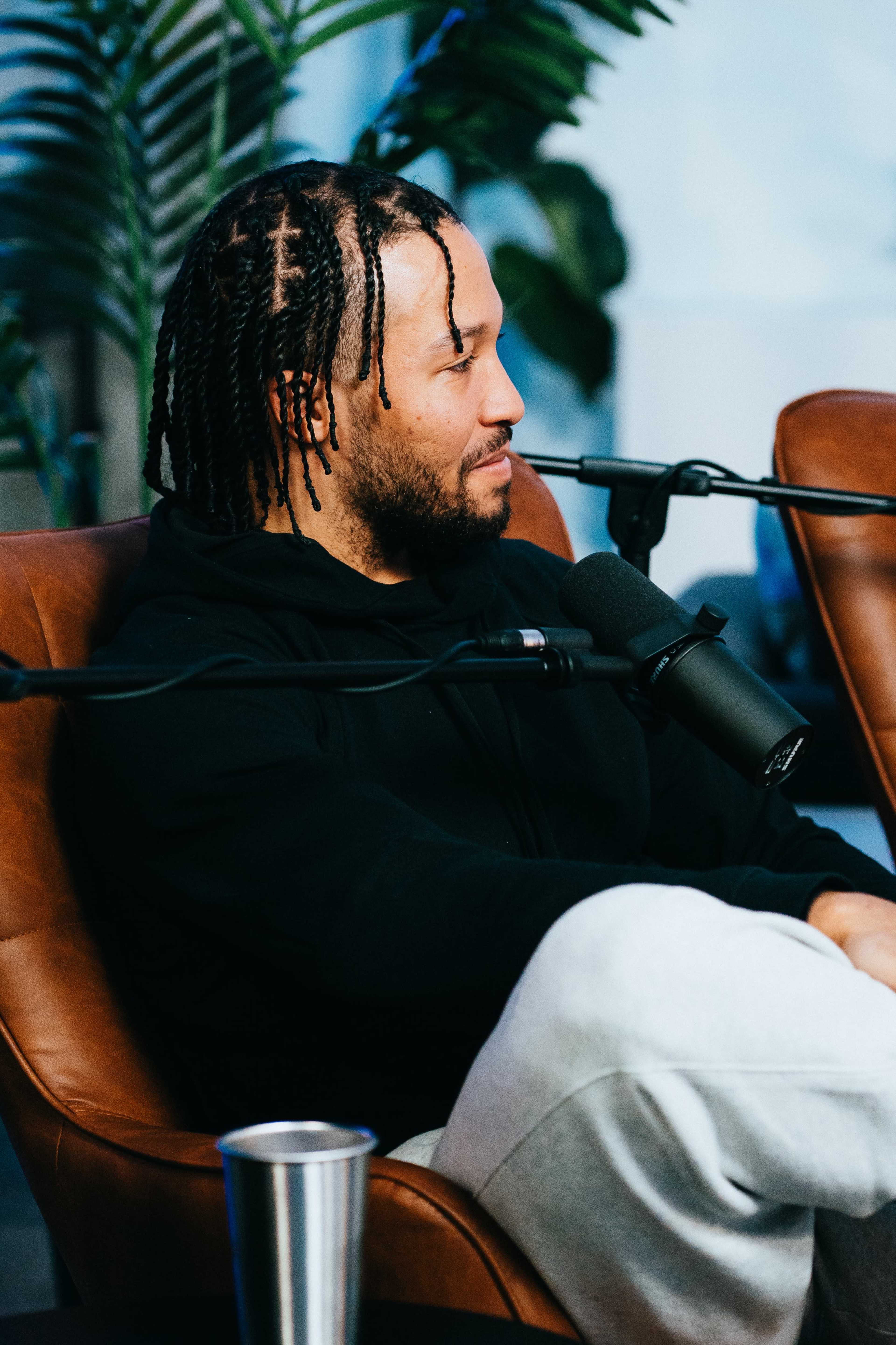 A man with braided hair sits in a brown leather chair, speaking into a microphone while wearing a black hoodie and gray sweatpants.