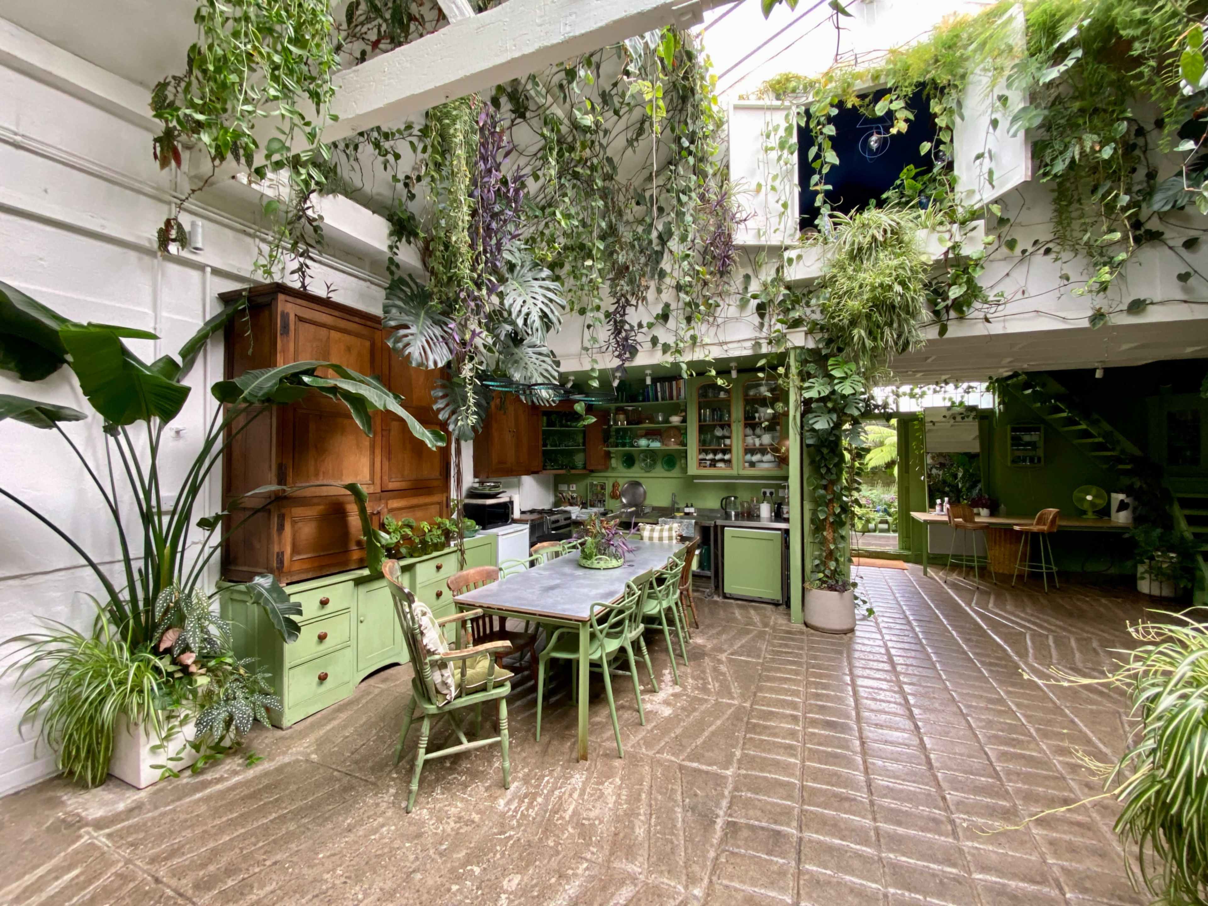 The spacious kitchen features green cabinetry, a large dining table, and abundant plants hanging from the ceiling and walls.