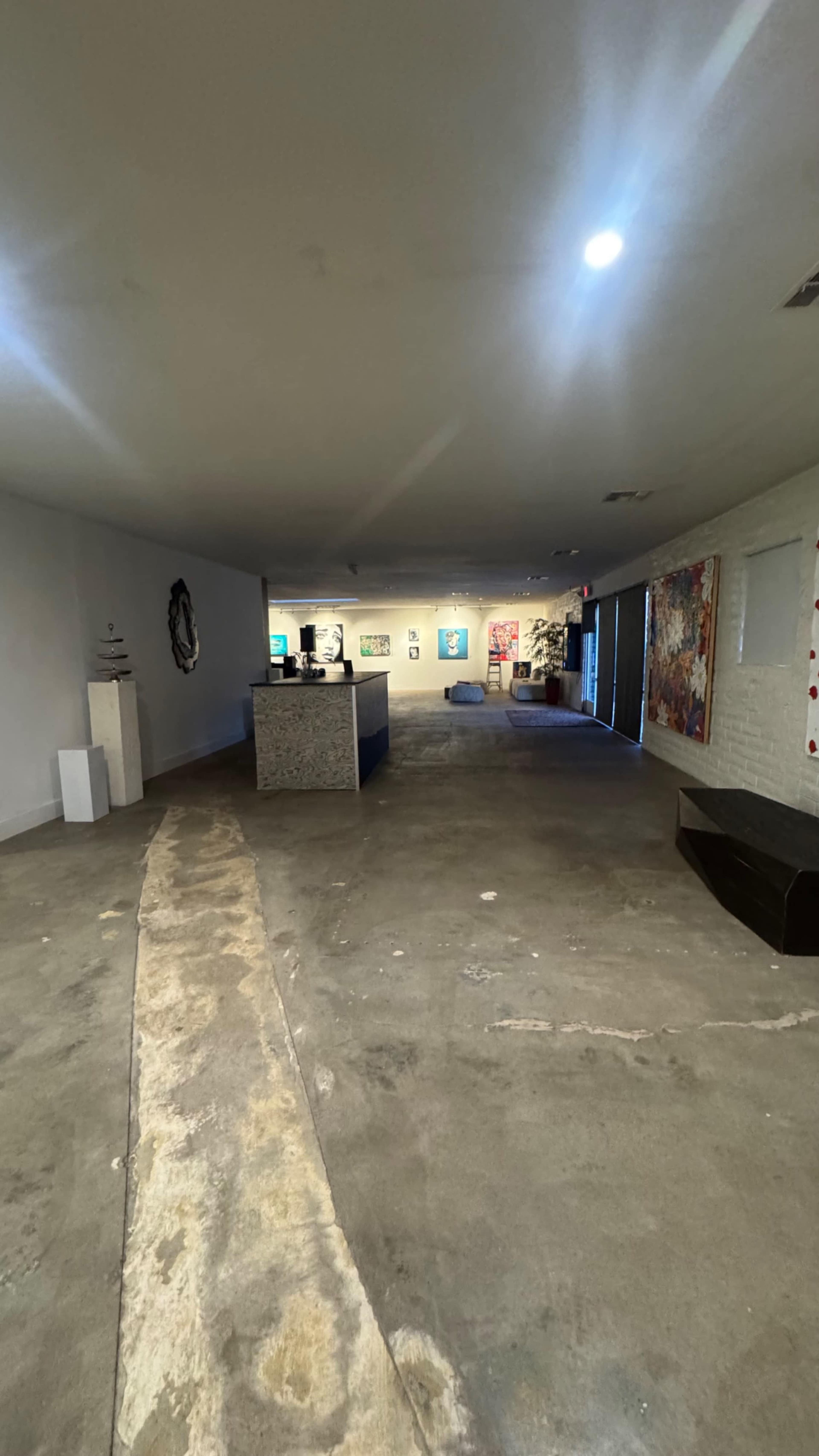 The image shows an interior gallery space with a concrete floor, artwork displayed on the walls, and a reception area in the center.