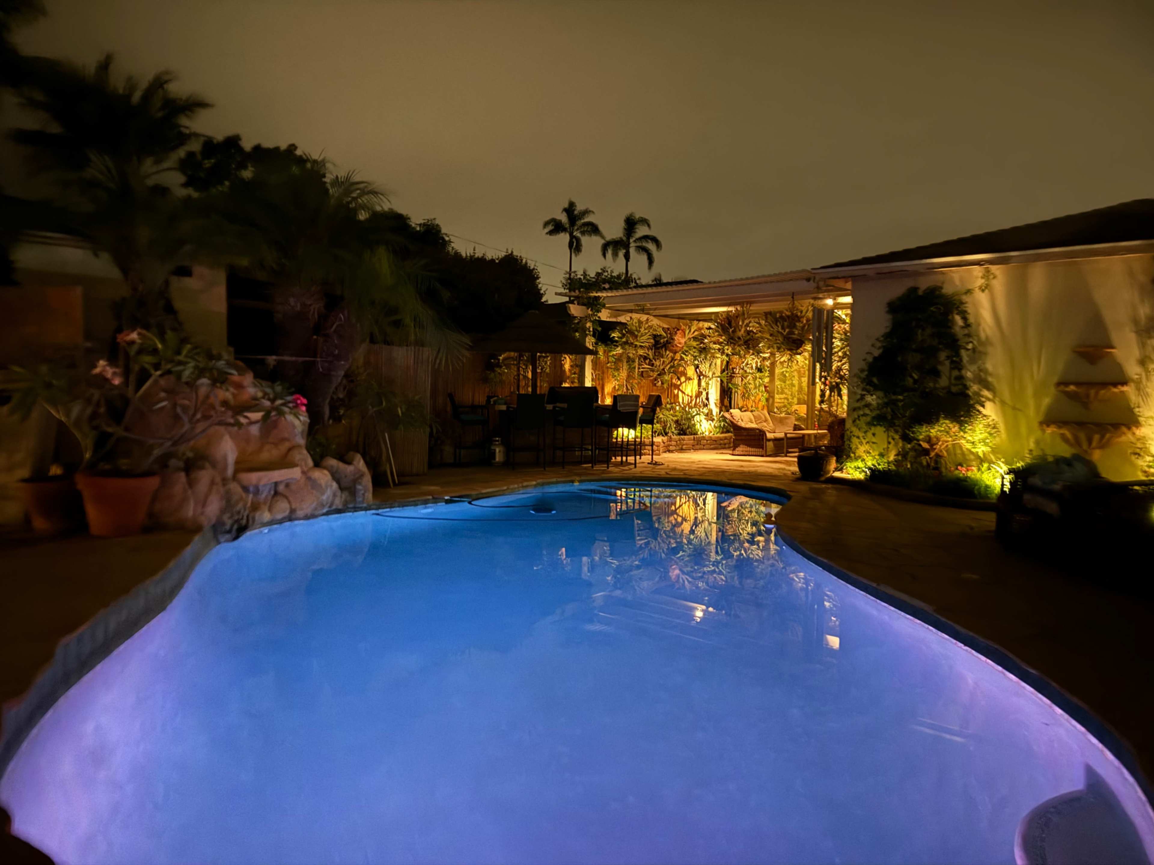A illuminated swimming pool with surrounding palm trees and a patio area featuring seating and plants under a night sky.