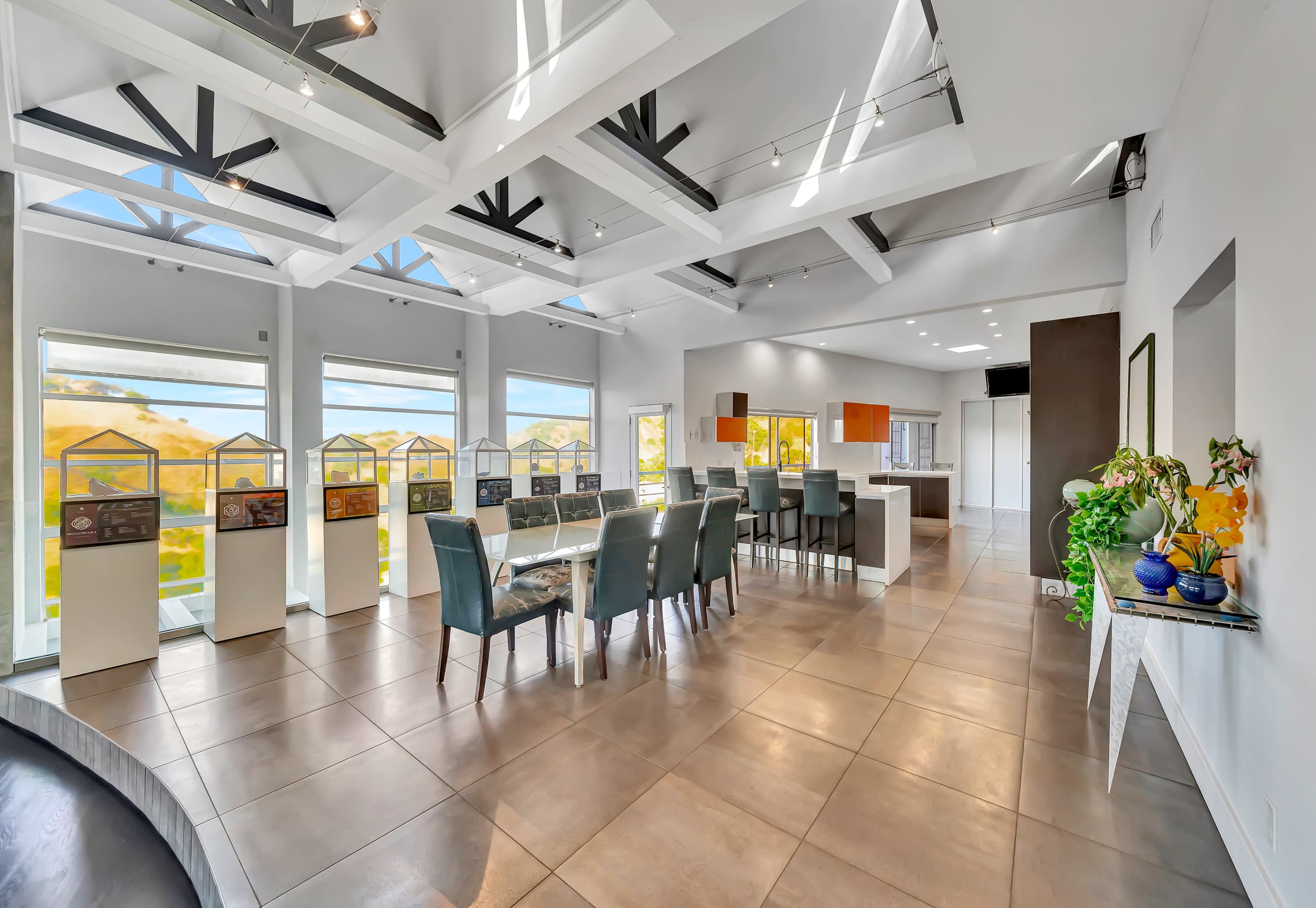 A spacious interior room features a dining area with a long table and chairs, large windows allowing natural light, and a modern kitchen in the background.