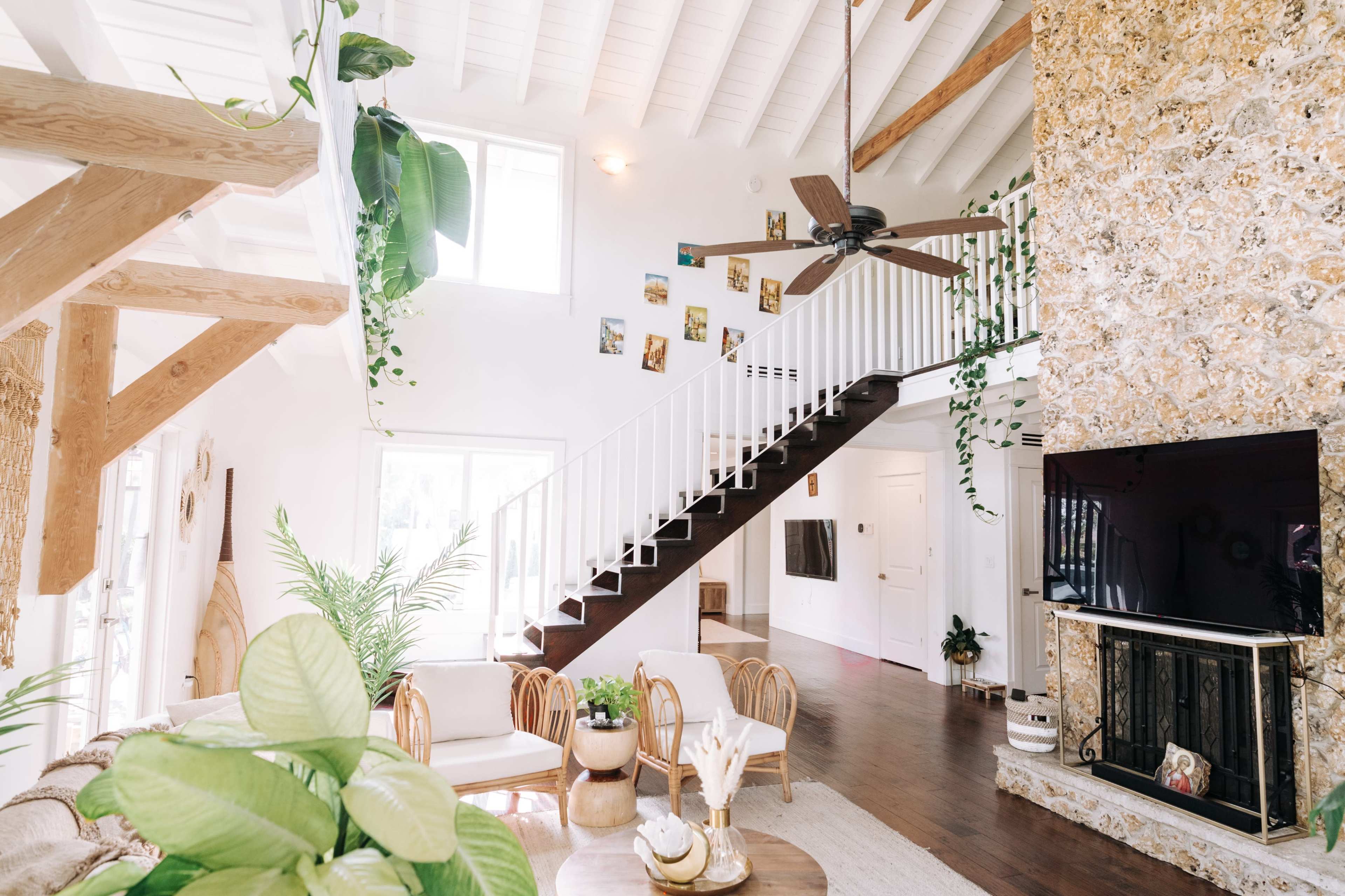 A bright living room features a staircase with white railings, a stone wall, and furniture arranged around a circular table.