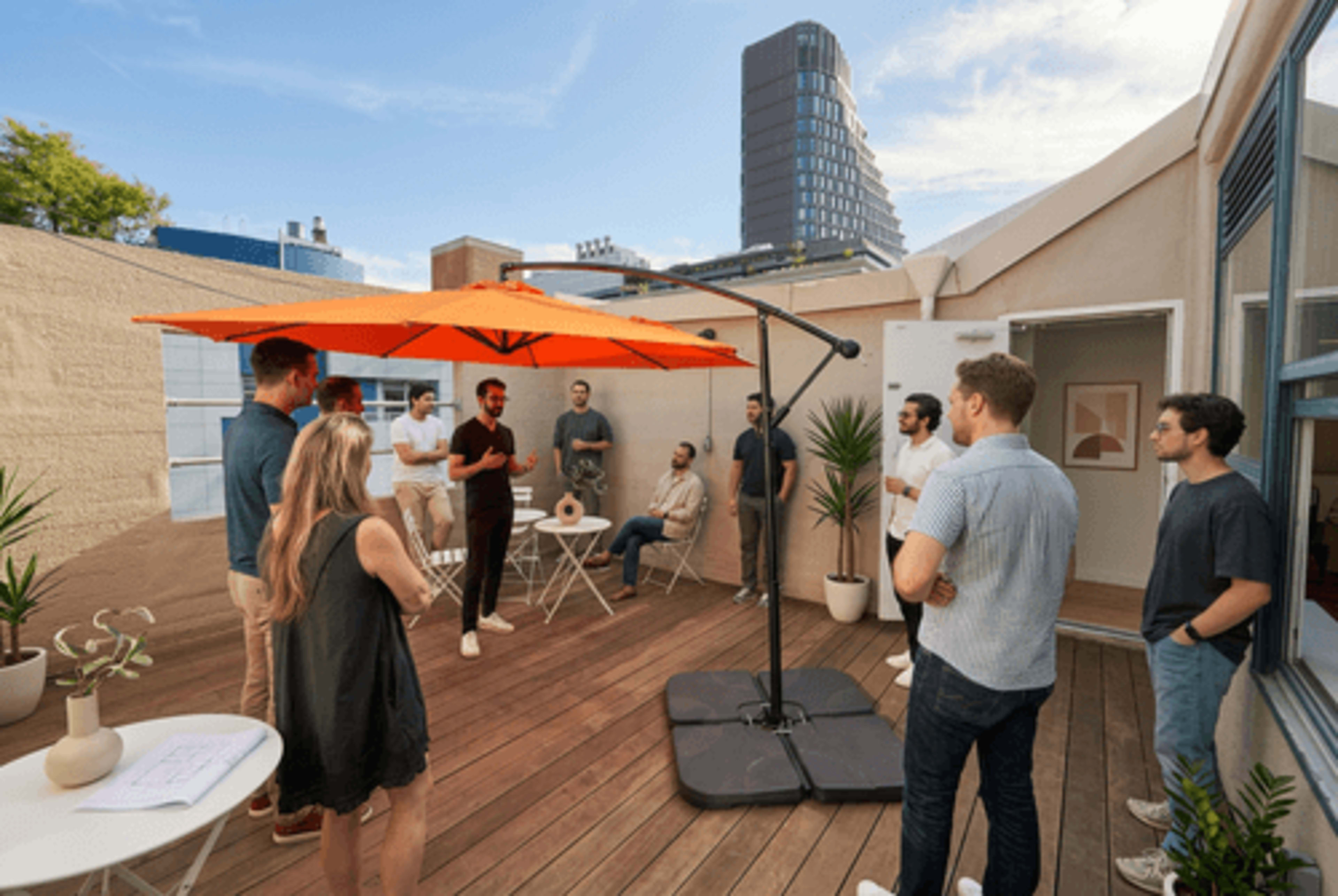 A group of people is gathered on a wooden terrace under an orange umbrella, engaging in discussion while surrounded by plants and modern furniture.