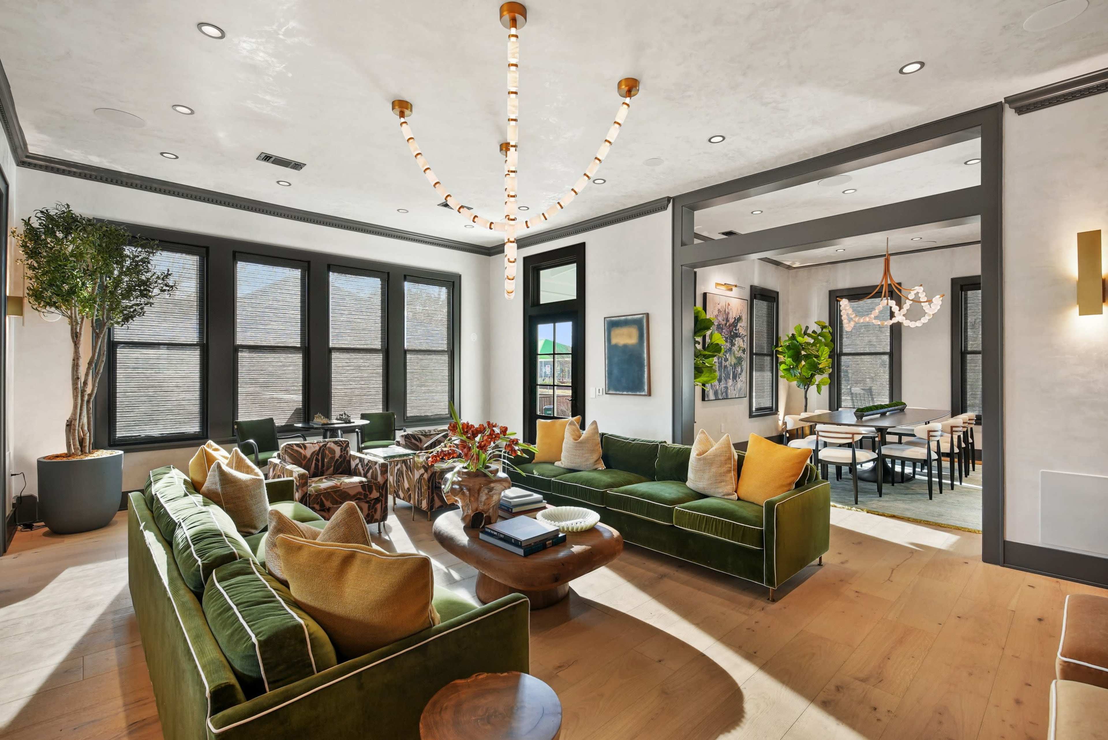 The image shows a modern living room with a green sectional sofa, a coffee table, and decorative plants, featuring large windows and a contemporary chandelier.