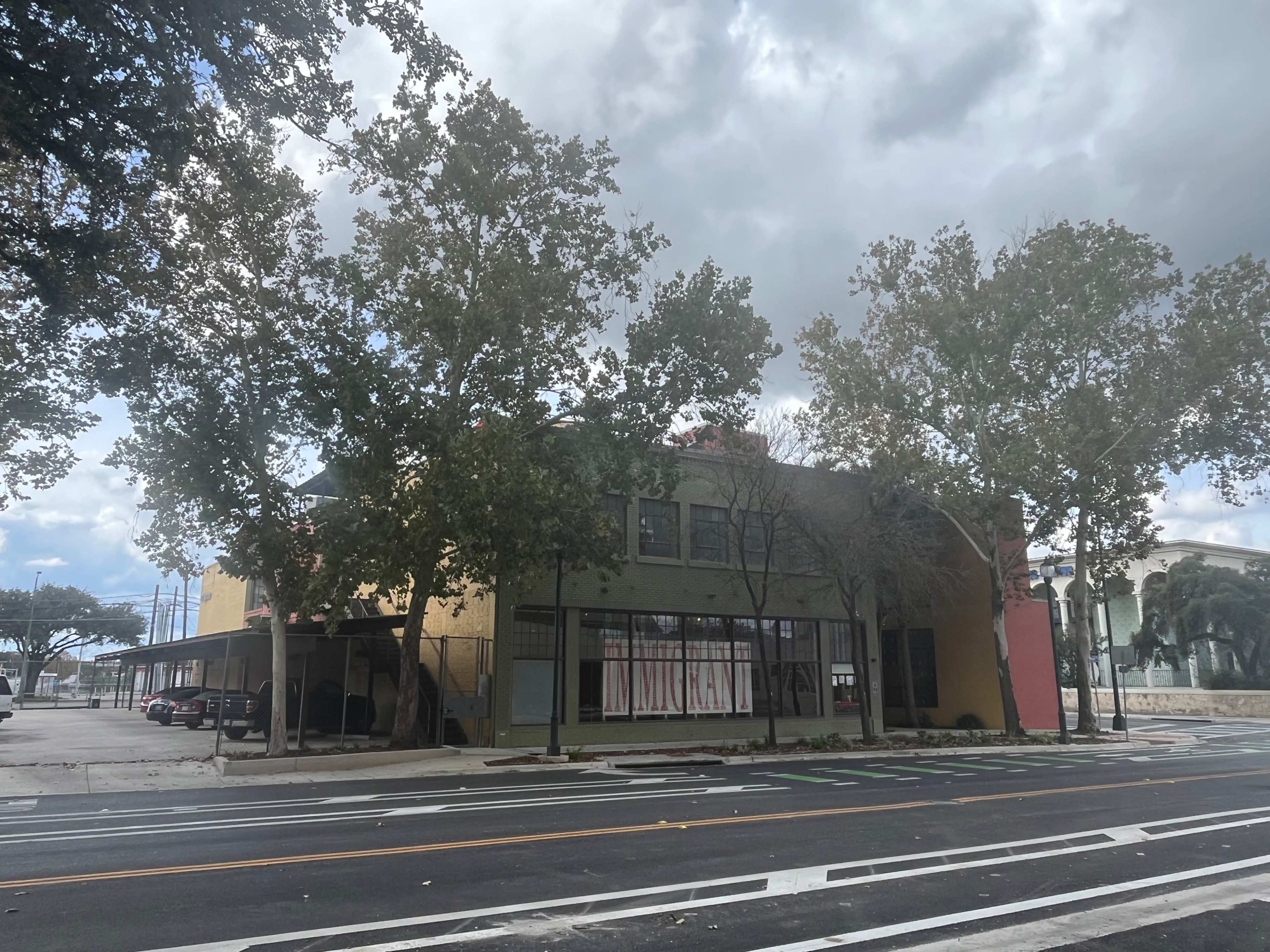 A two-story building with a green facade and large windows is flanked by several trees along a street under a cloudy sky.
