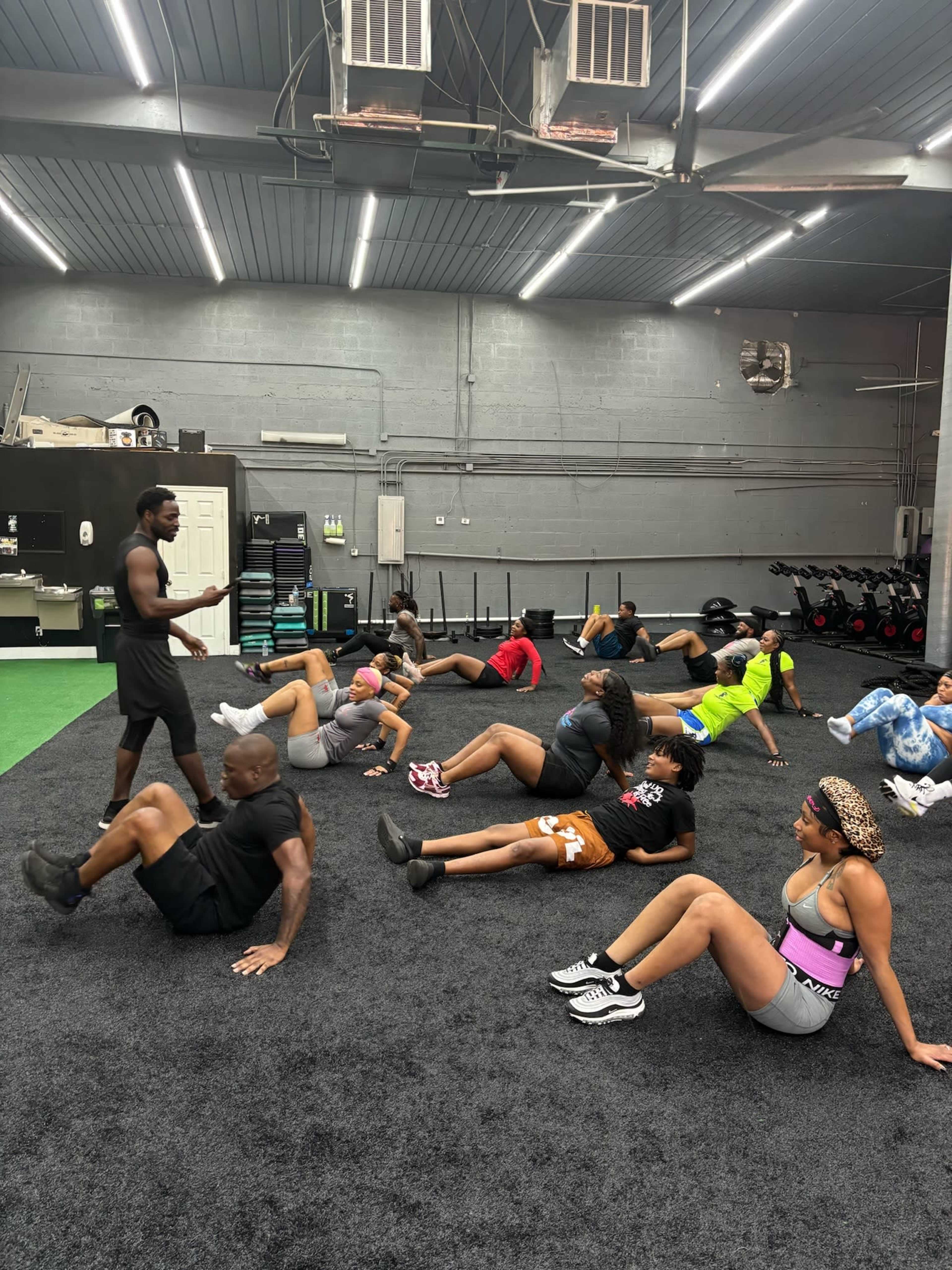 A fitness instructor guides a group of individuals performing sit-up exercises on a black carpeted gym floor.