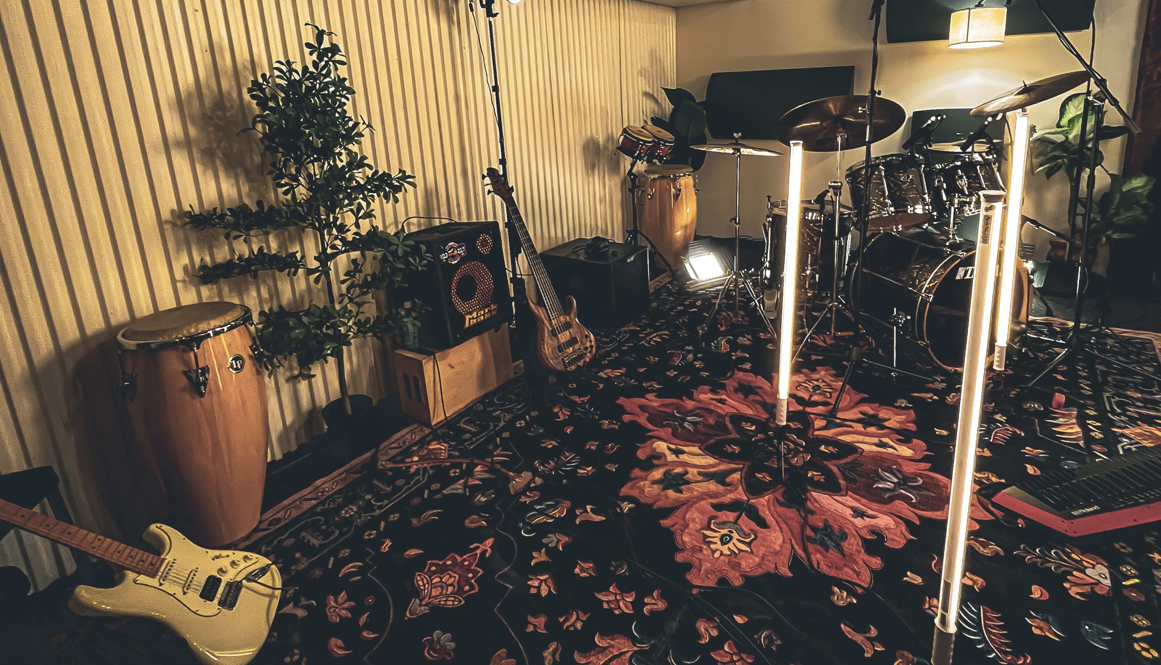 The image shows a music rehearsal space featuring various instruments, including a guitar, congas, a bass guitar, and a drum set, arranged on a patterned rug.