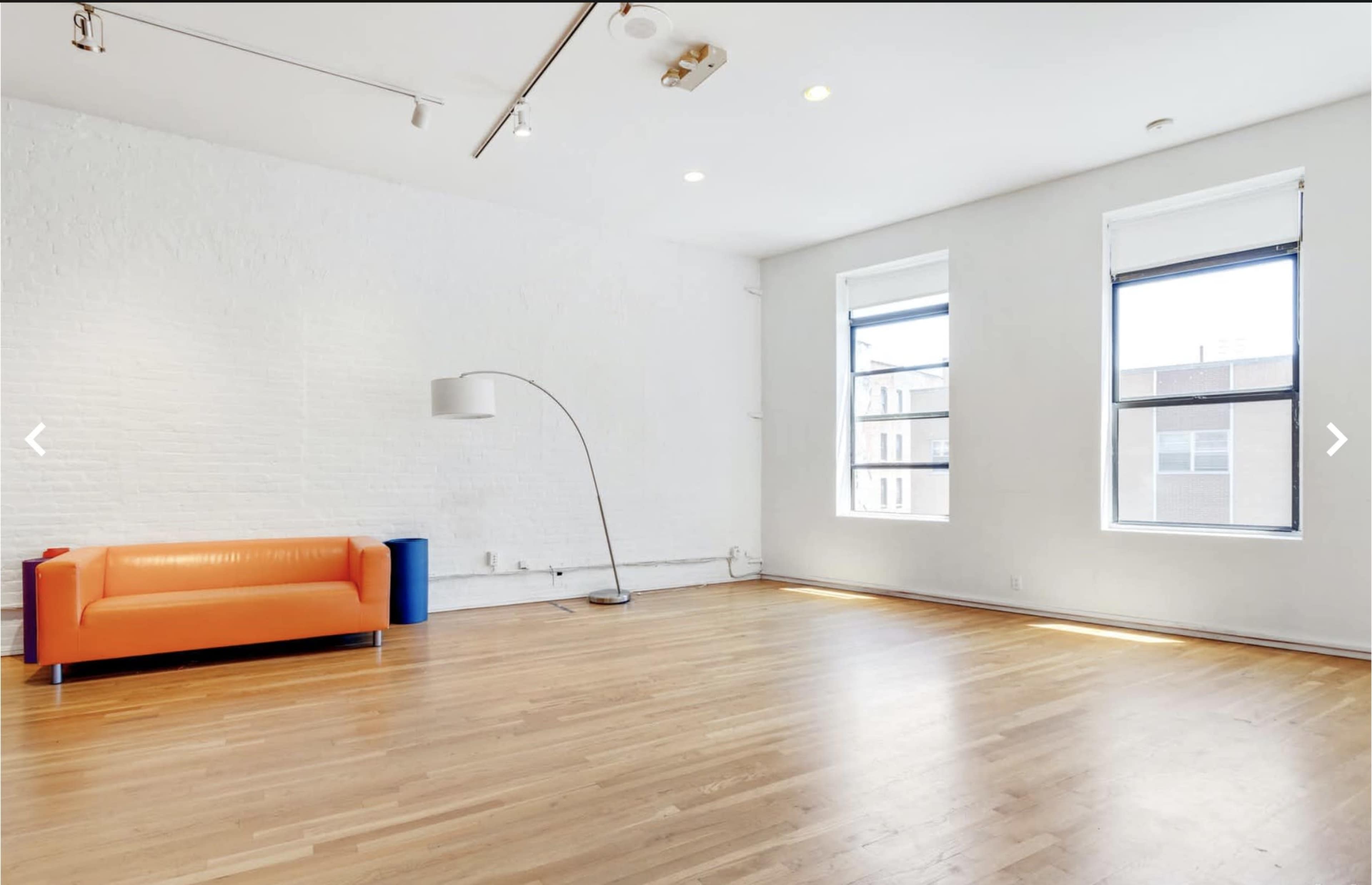 A spacious room with two windows, a modern orange couch, a standing lamp, and a blue cylinder beside it on a hardwood floor.