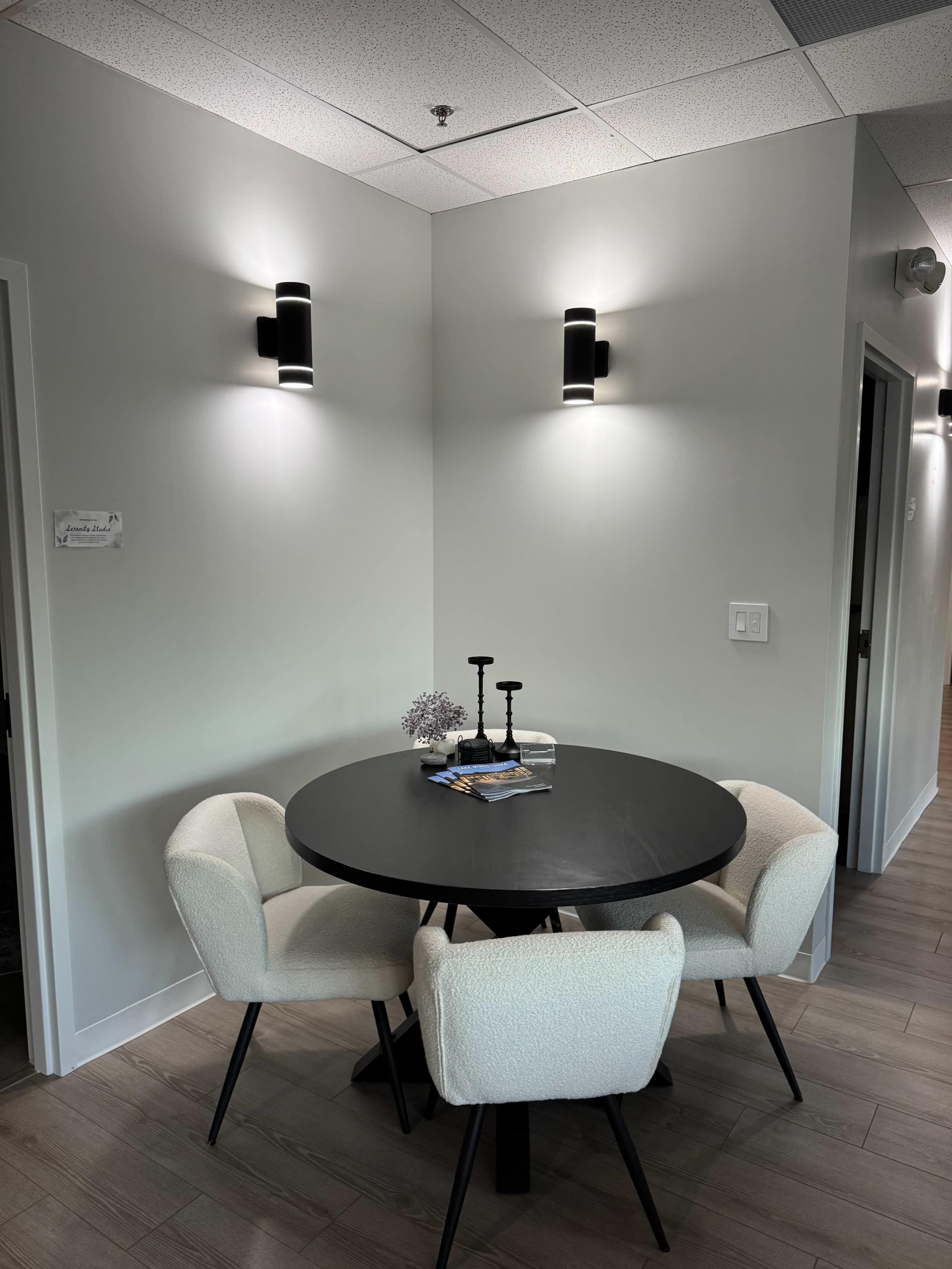 A circular black table with two white chairs is situated in a well-lit corner of a room with modern lighting fixtures on the walls.