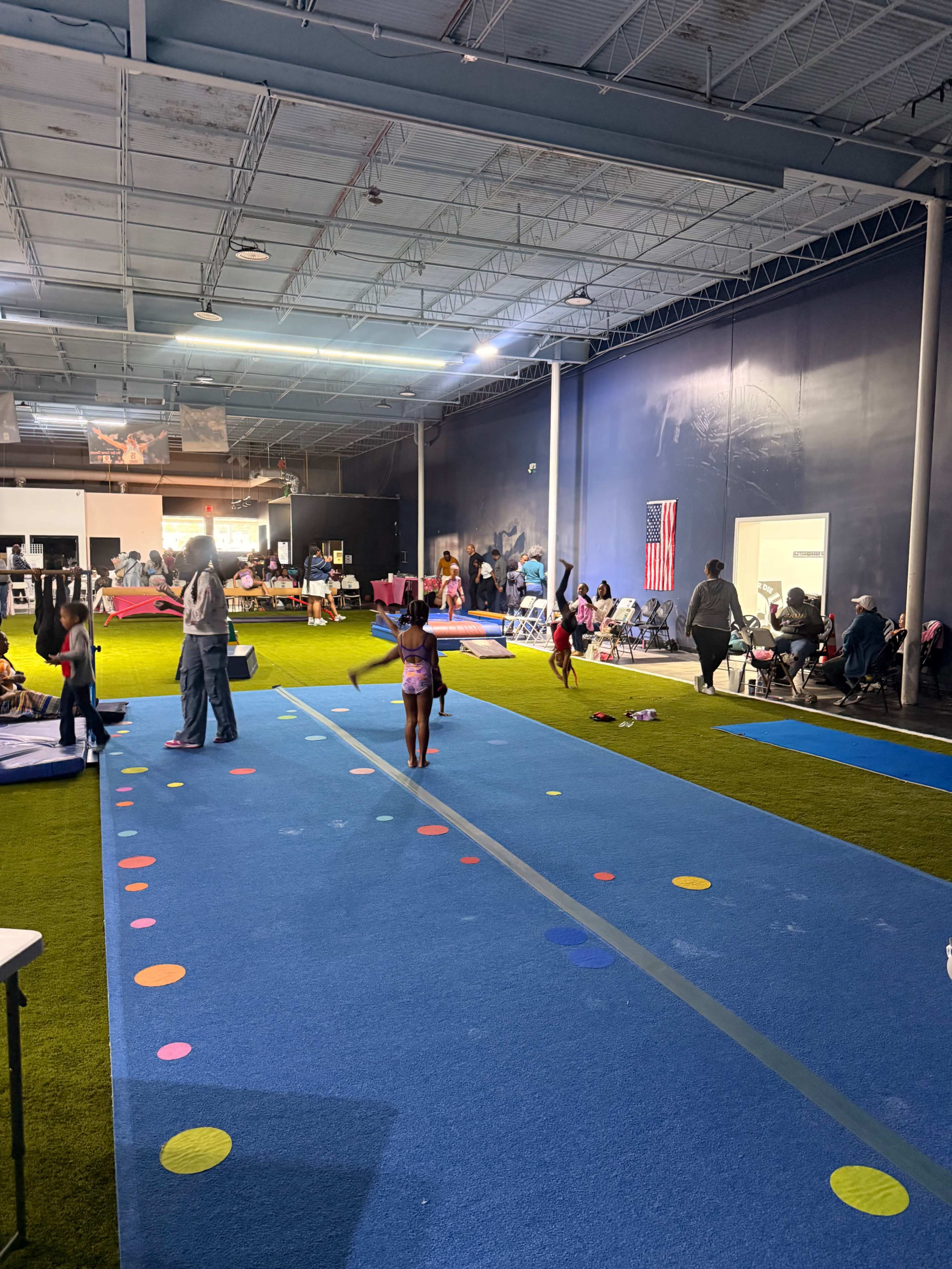 A group of children practices gymnastics on a colorful mat in a spacious indoor facility while adults observe from seating areas.