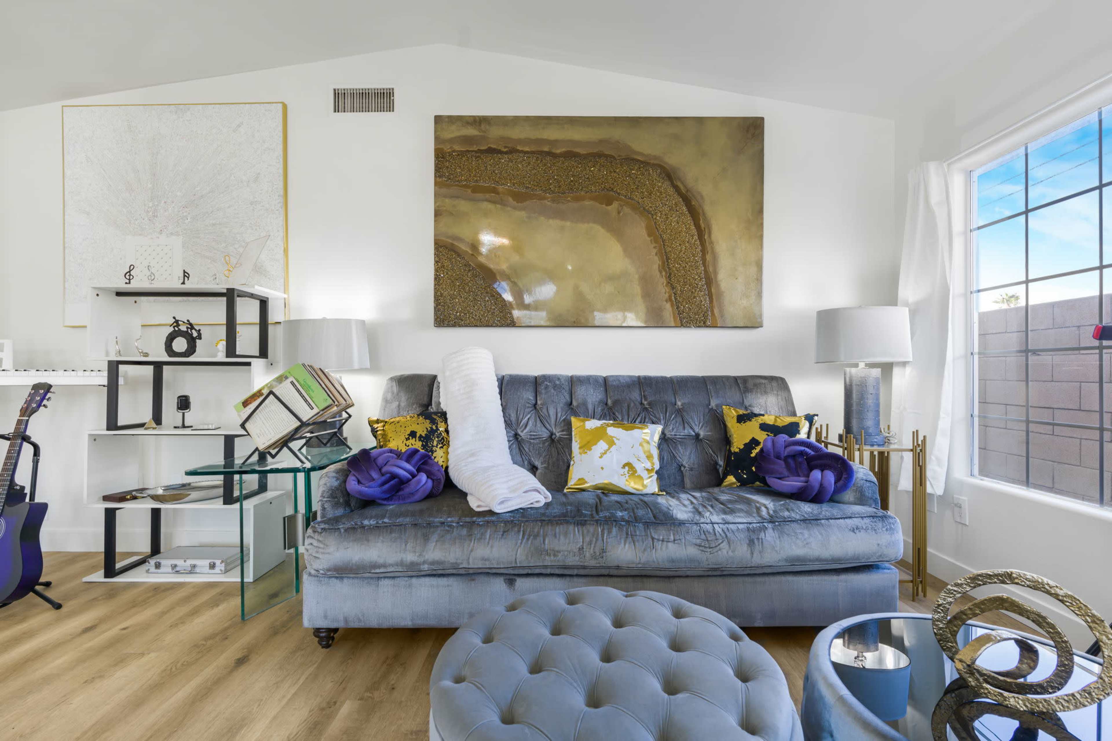 A modern living room features a gray sofa with decorative pillows, a glass side table, and artwork on the walls, illuminated by natural light from a nearby window.