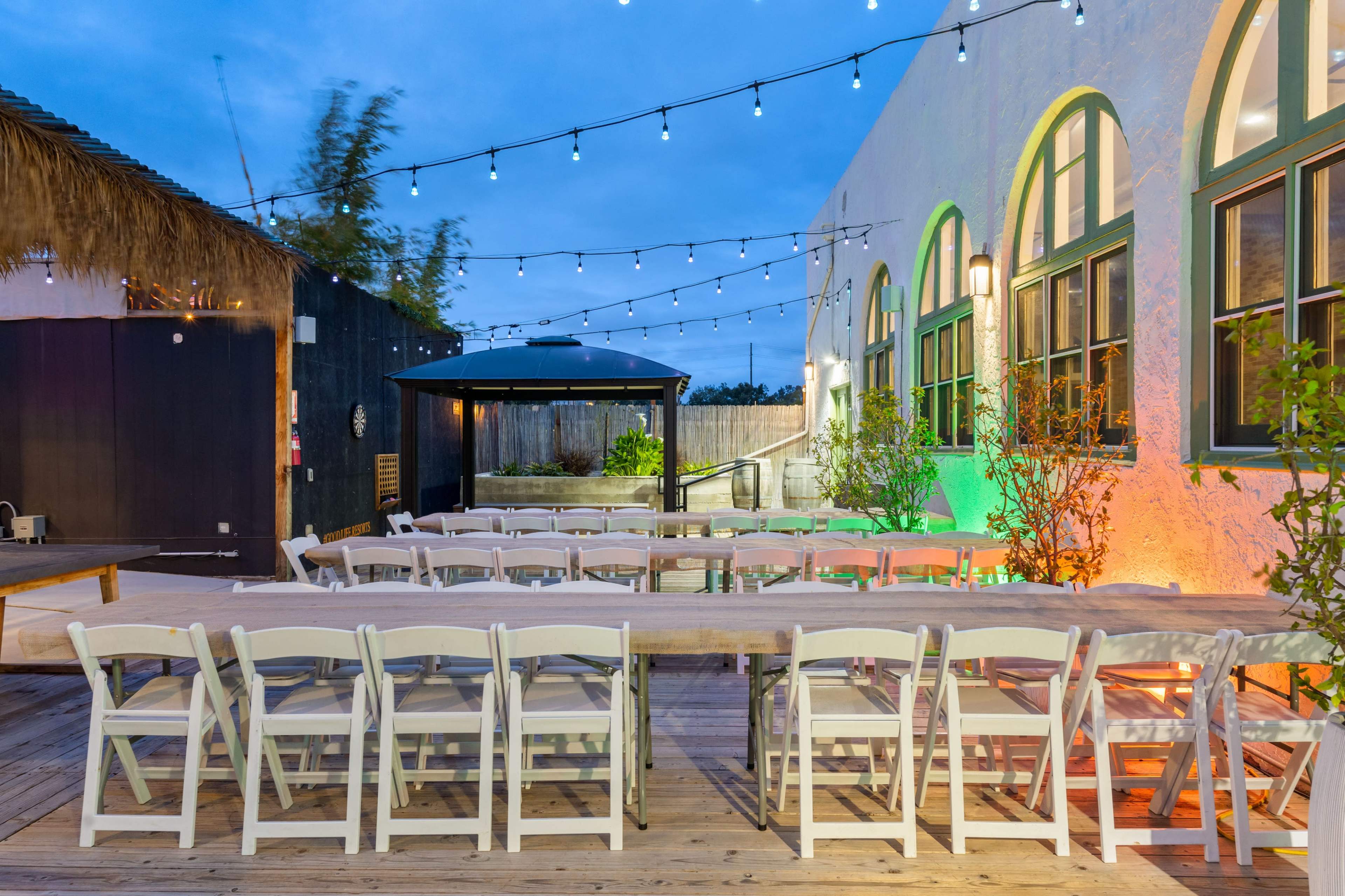 An outdoor dining area is set up with a long wooden table surrounded by white chairs, illuminated by string lights against a backdrop of a well-lit building.