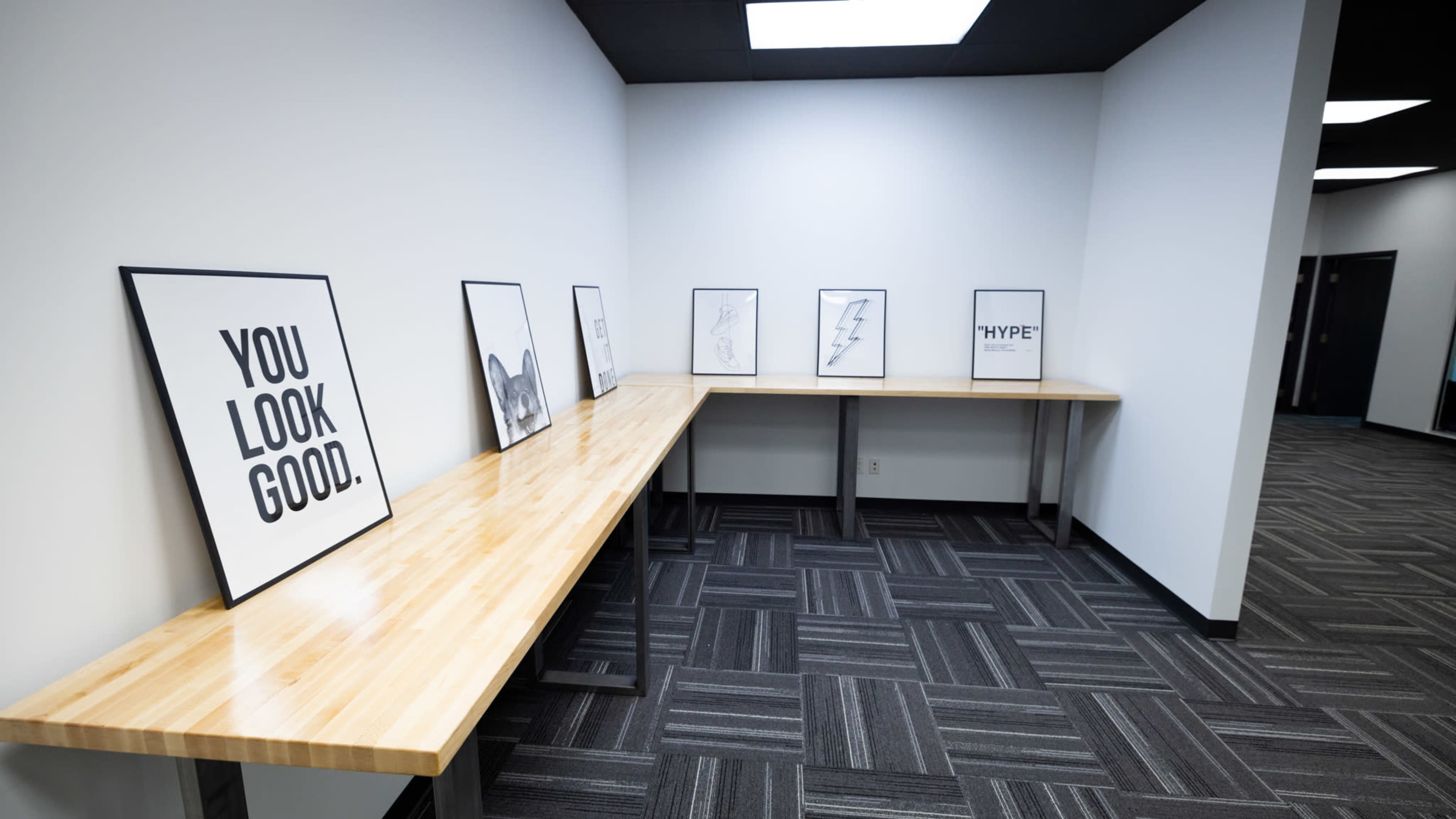 A minimalistic room features a long wooden table against a wall, displaying various framed posters with bold black text and designs.