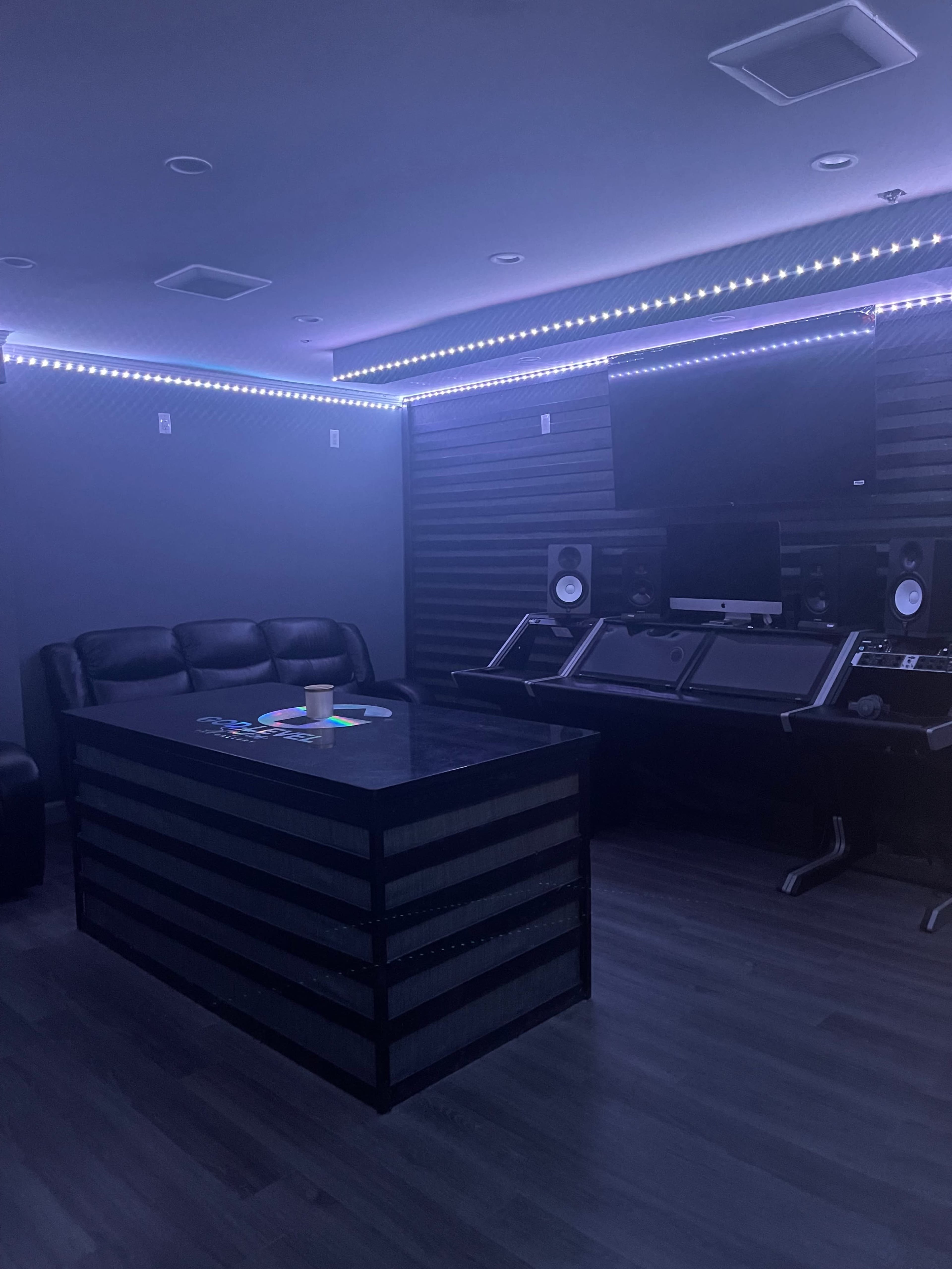 A dimly lit music studio features a black table at the center, surrounded by sound equipment, with a couch and LED lights along the walls.