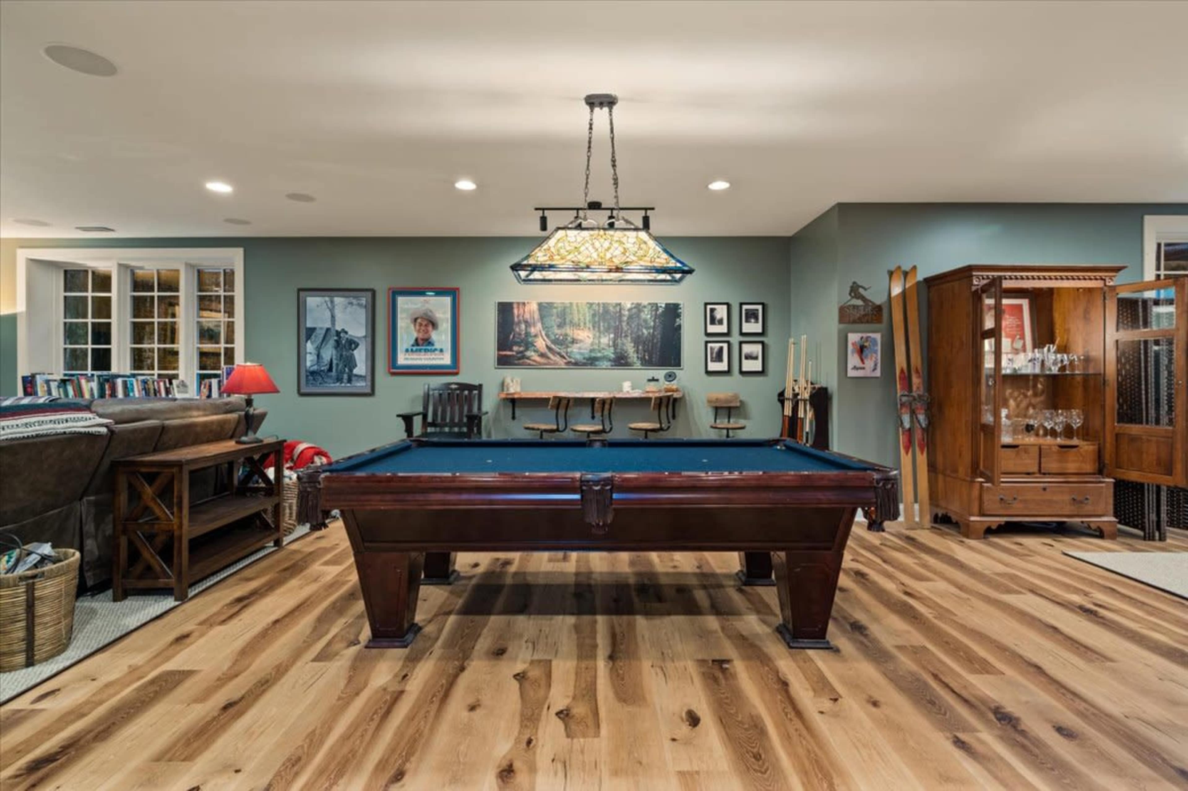 A billiards table is centered in a recreational room featuring wooden flooring, various artworks on the walls, and furniture arranged around the space.