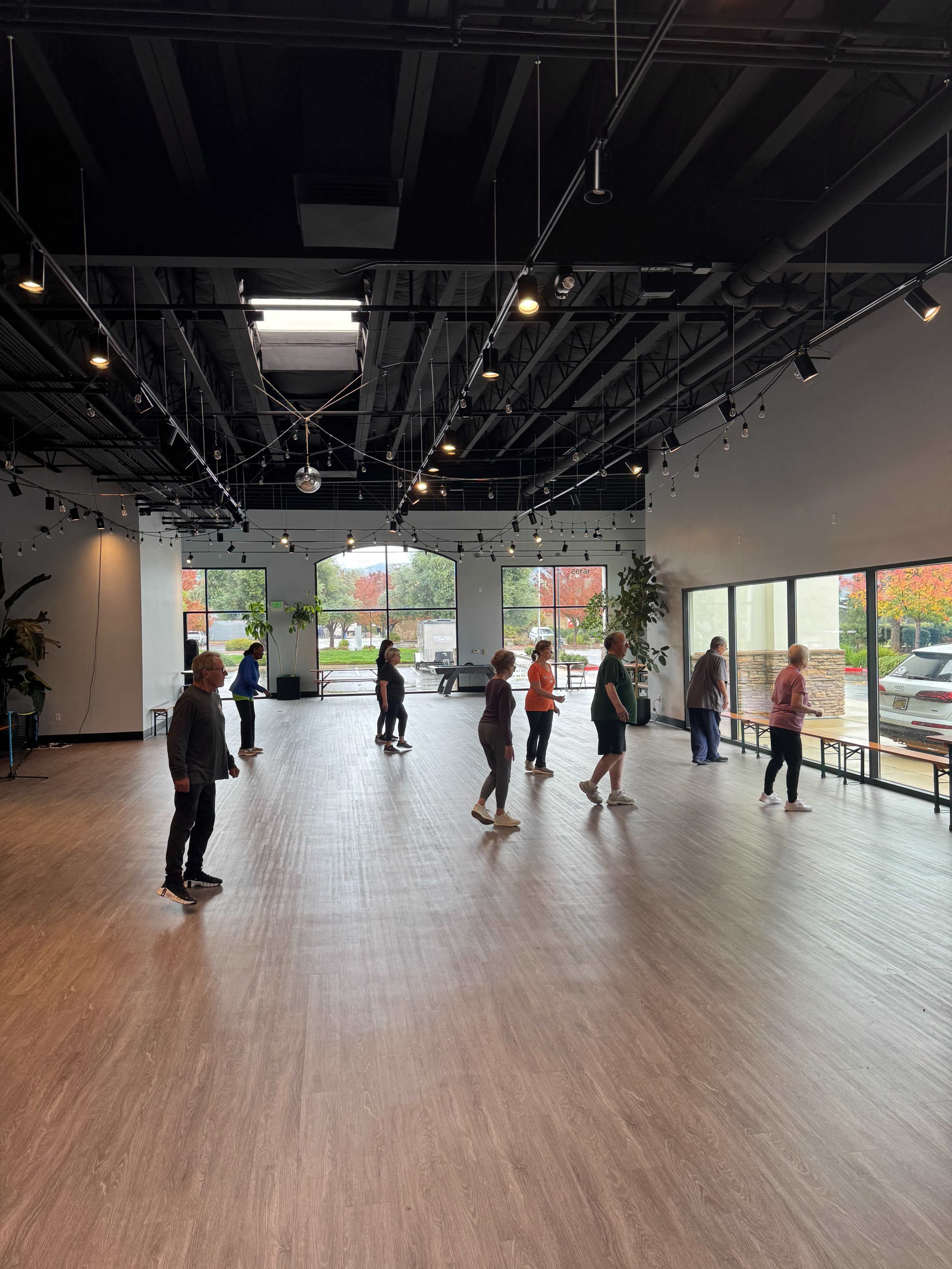 A group of people is dancing in a spacious studio with large windows and wooden flooring.