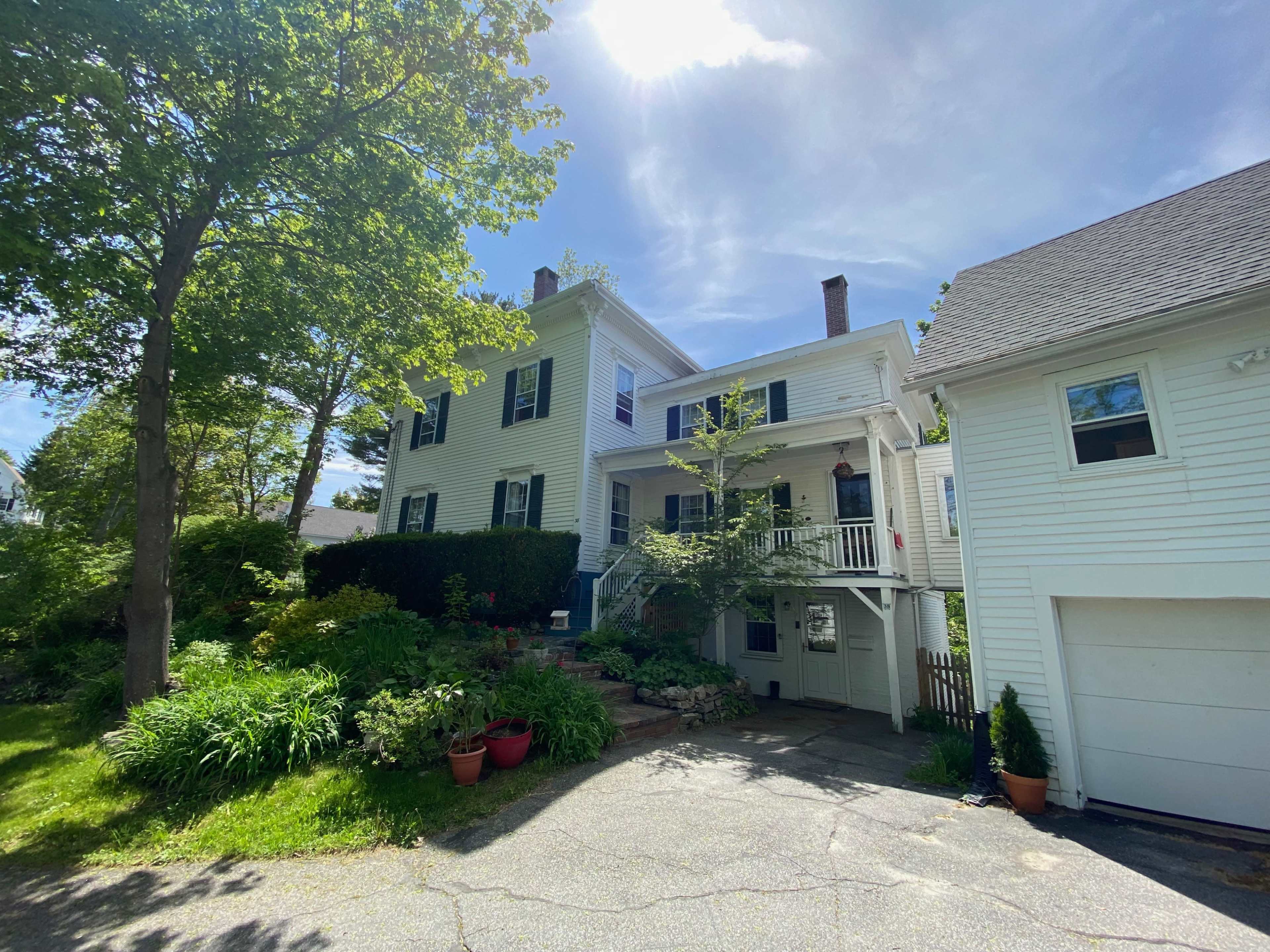 A two-story yellow house surrounded by greenery and flowers, with a visible garage on the right.