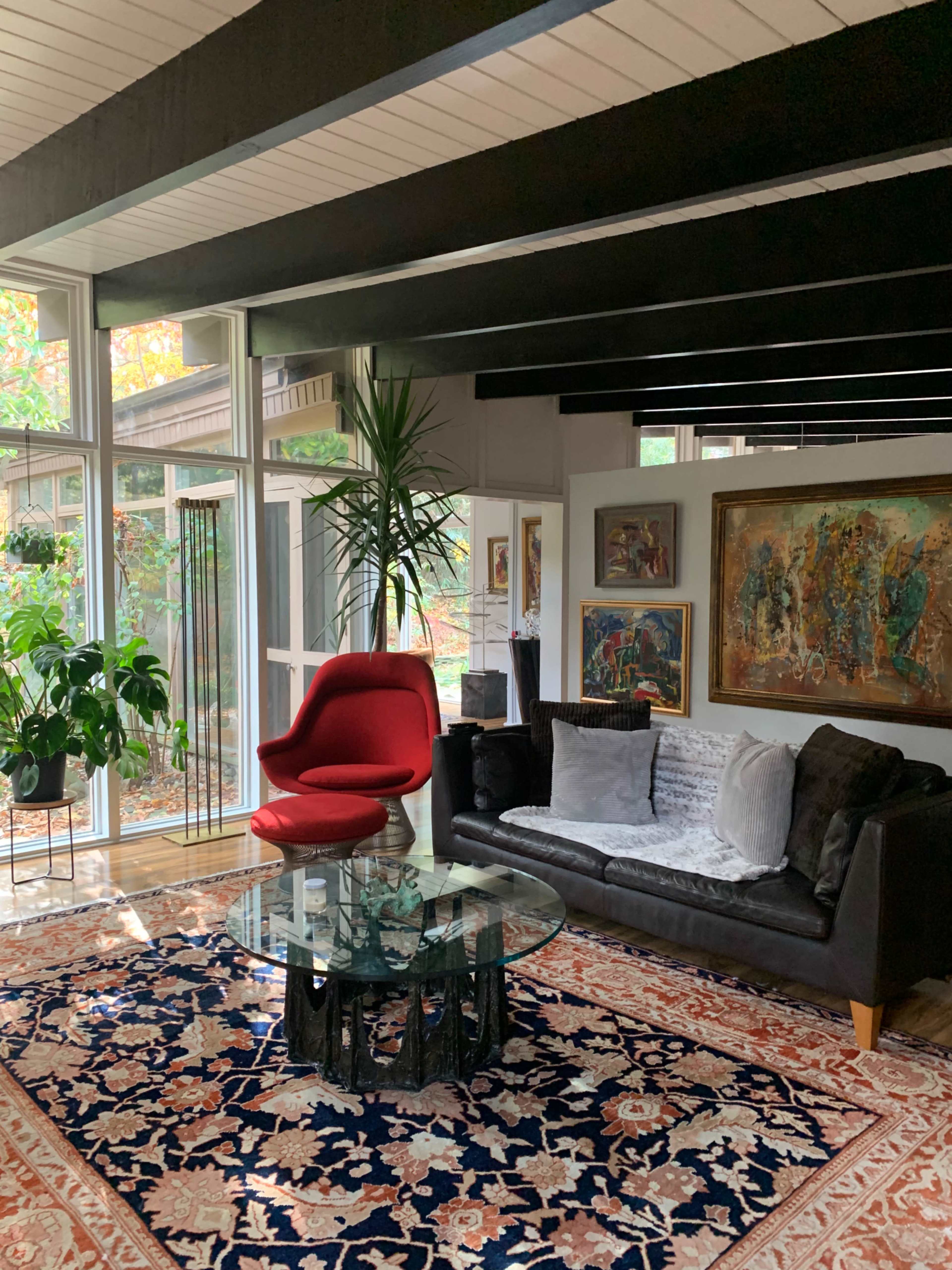A modern living room features a red accent chair, a black sofa, a glass coffee table, and various artworks against a backdrop of large windows.