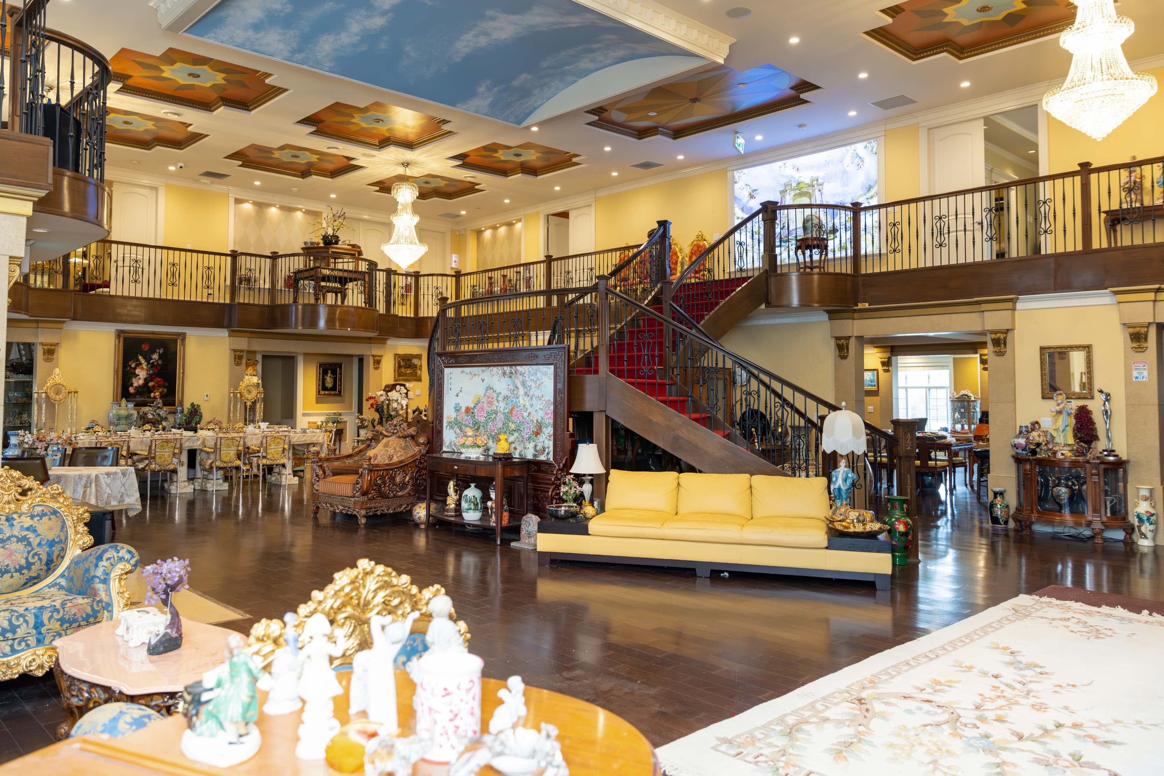 The image shows a spacious, elegantly furnished foyer with a high ceiling, featuring a large staircase, ornate furniture, and various decorative items throughout the room.