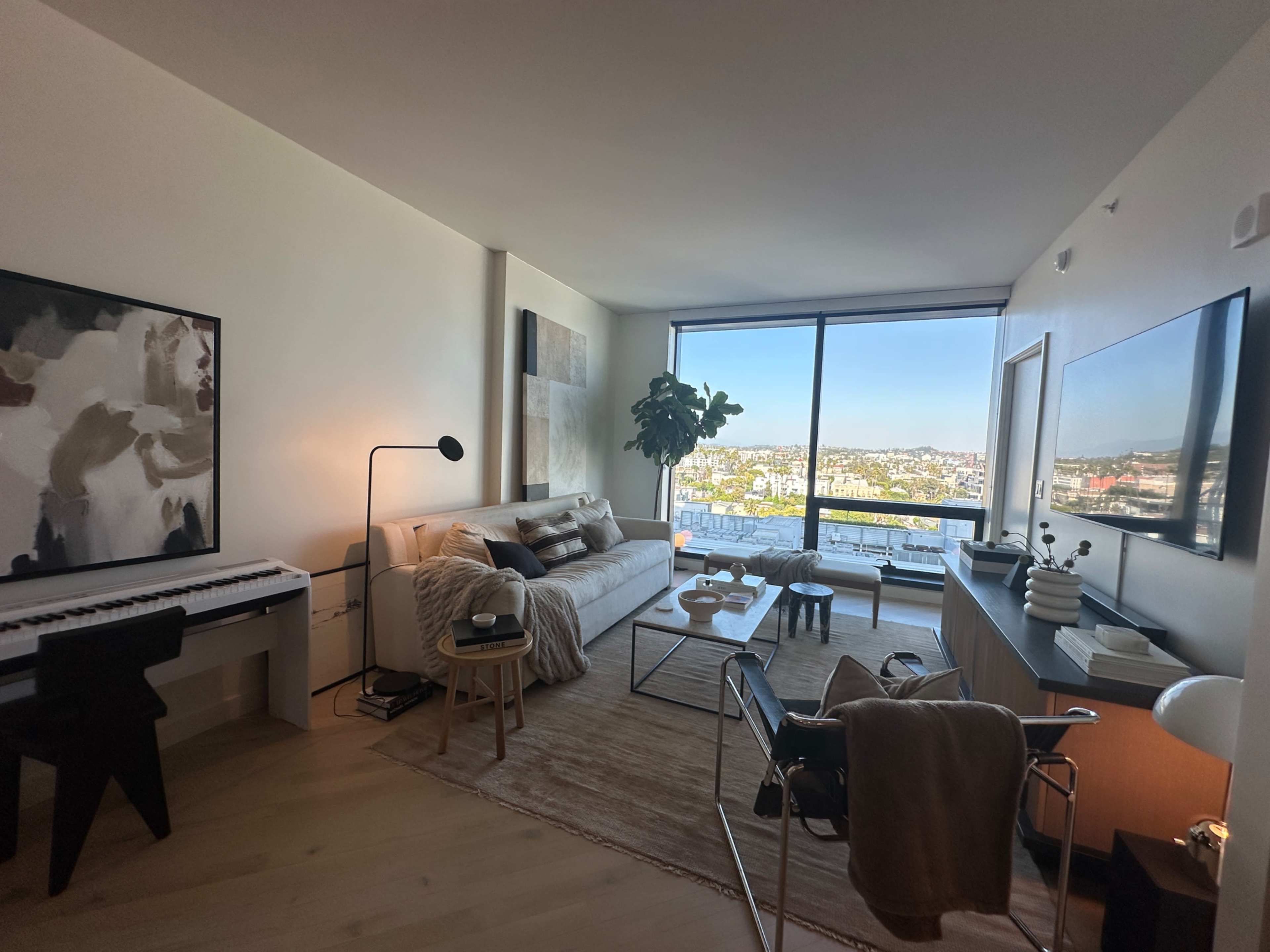 The image shows a modern living room with a sofa, coffee table, and piano, featuring large windows that overlook a cityscape.