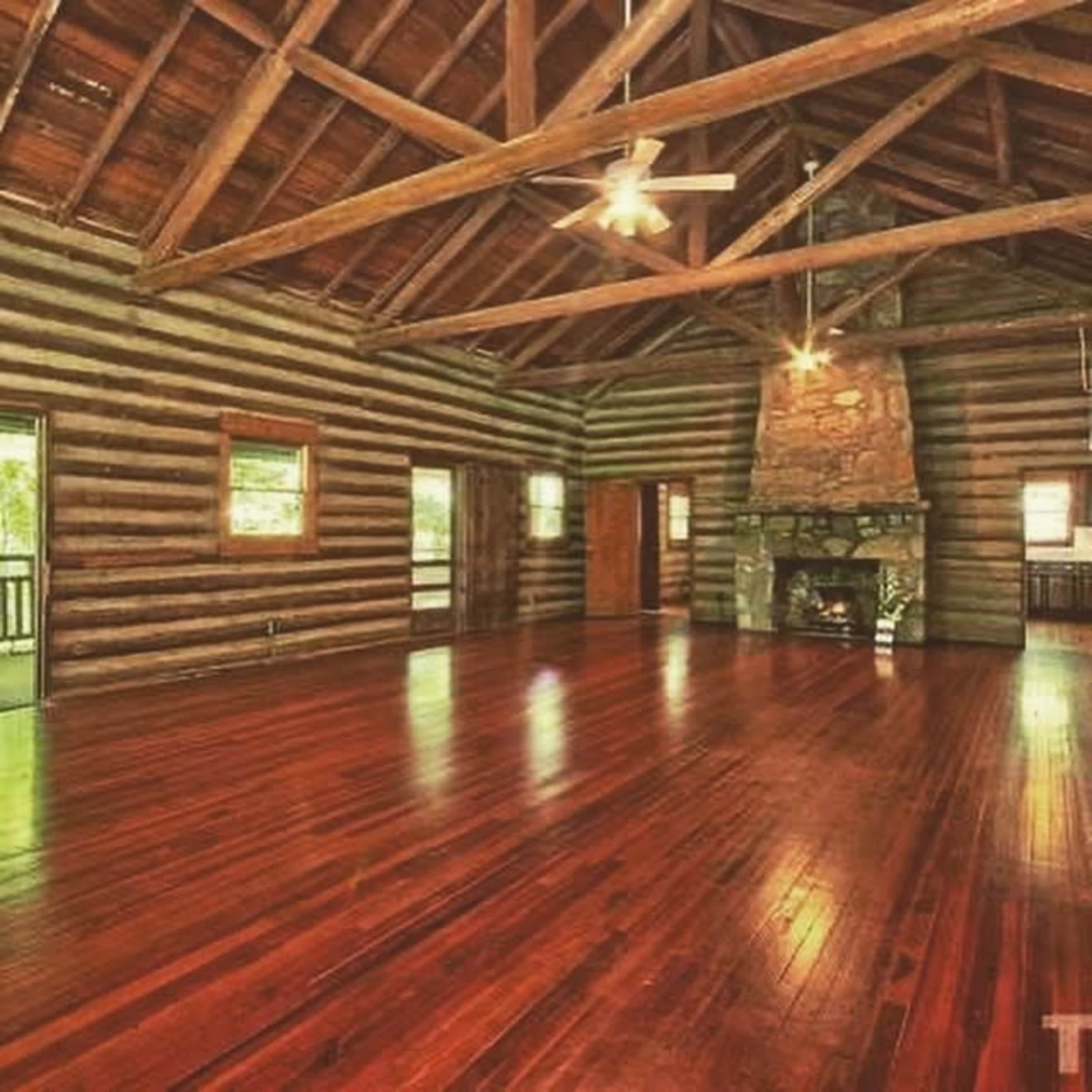 The image shows a spacious log cabin interior with wooden beams, a stone fireplace, and polished hardwood floors.
