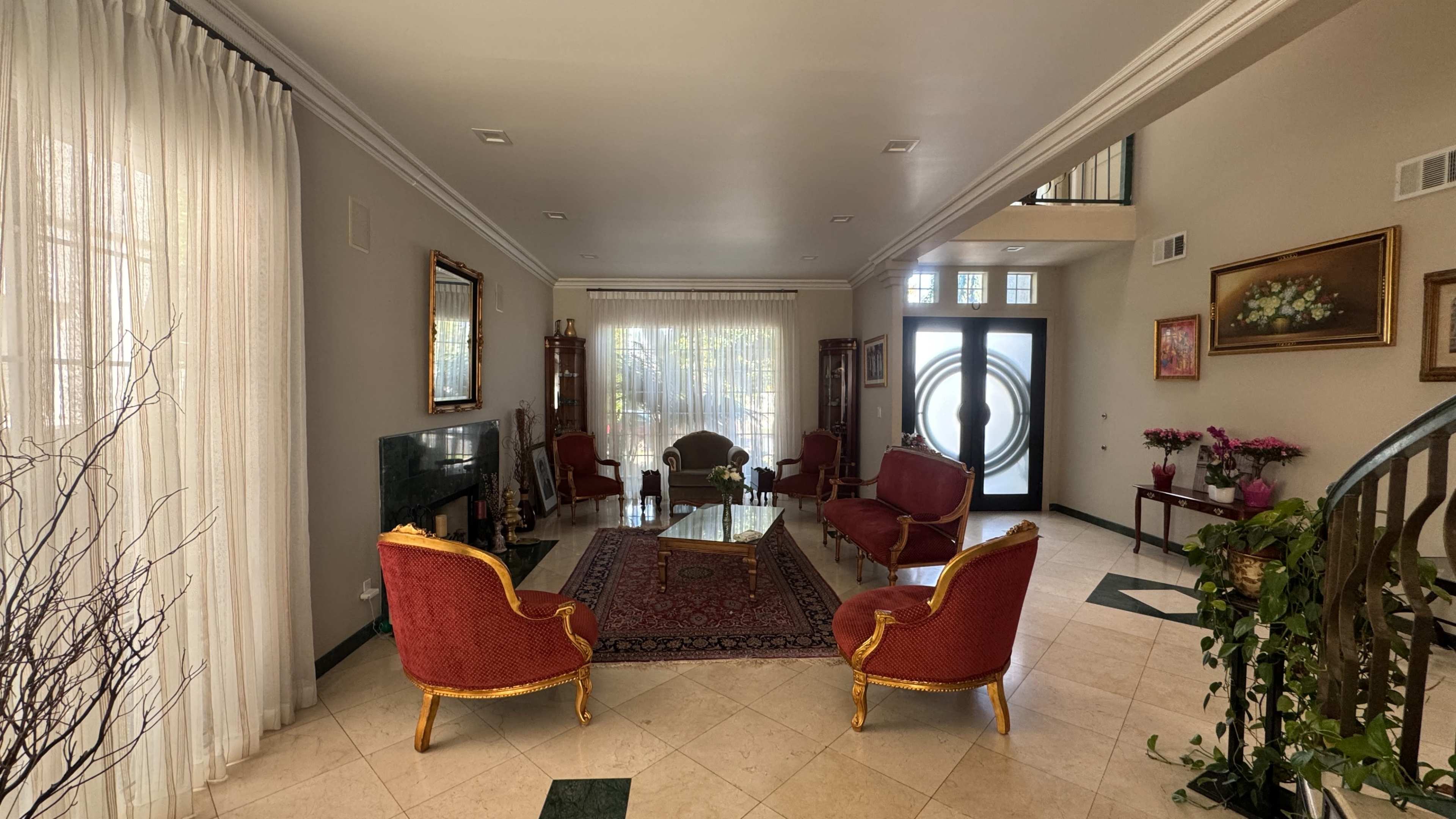 The image shows a spacious living room with four red armchairs arranged around a glass coffee table on a patterned rug, facing large windows with sheer curtains.