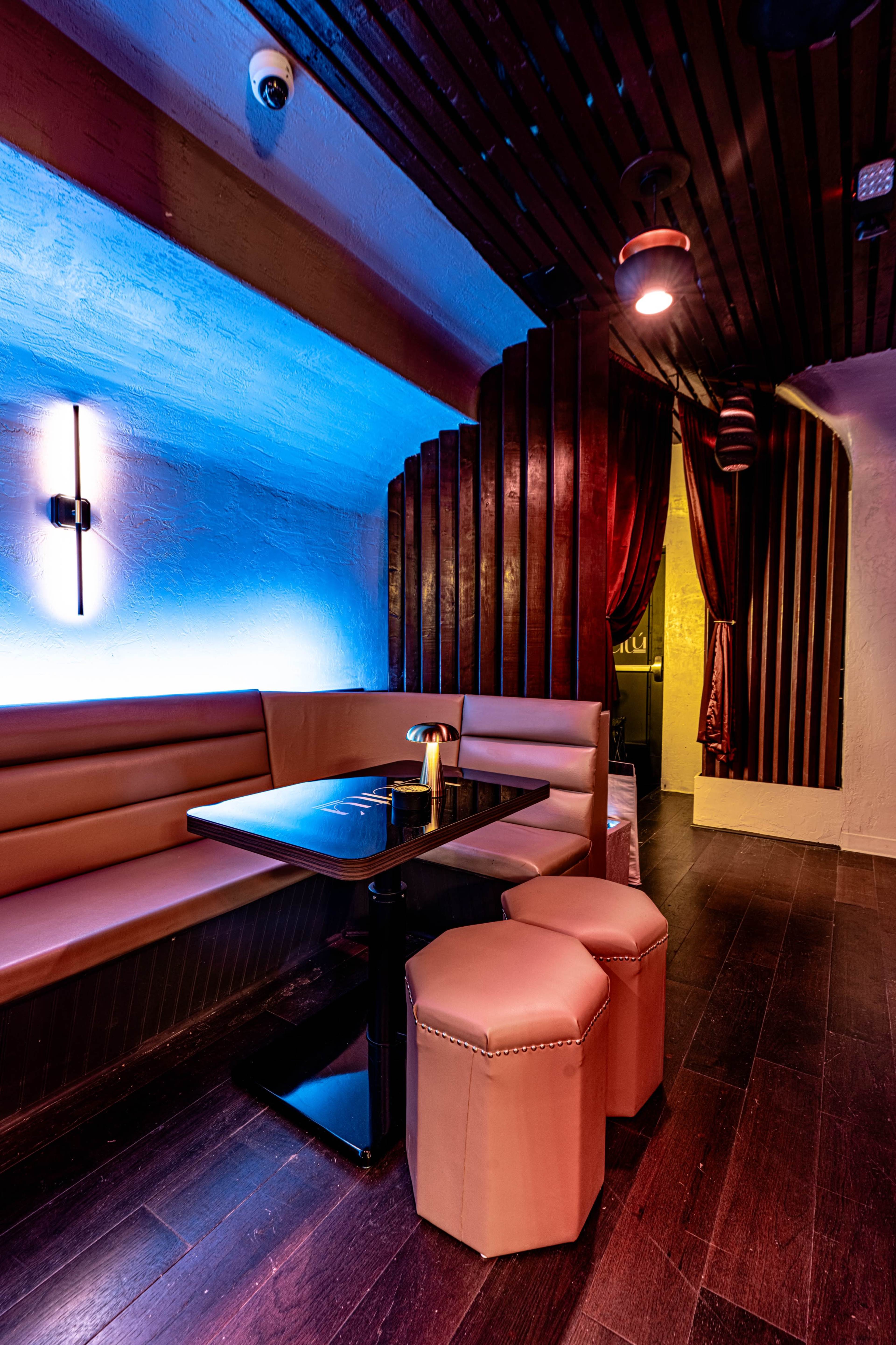 The image shows a dimly lit lounge area featuring a booth with a black table and two cylindrical stools, accented by blue lighting and wooden paneling.