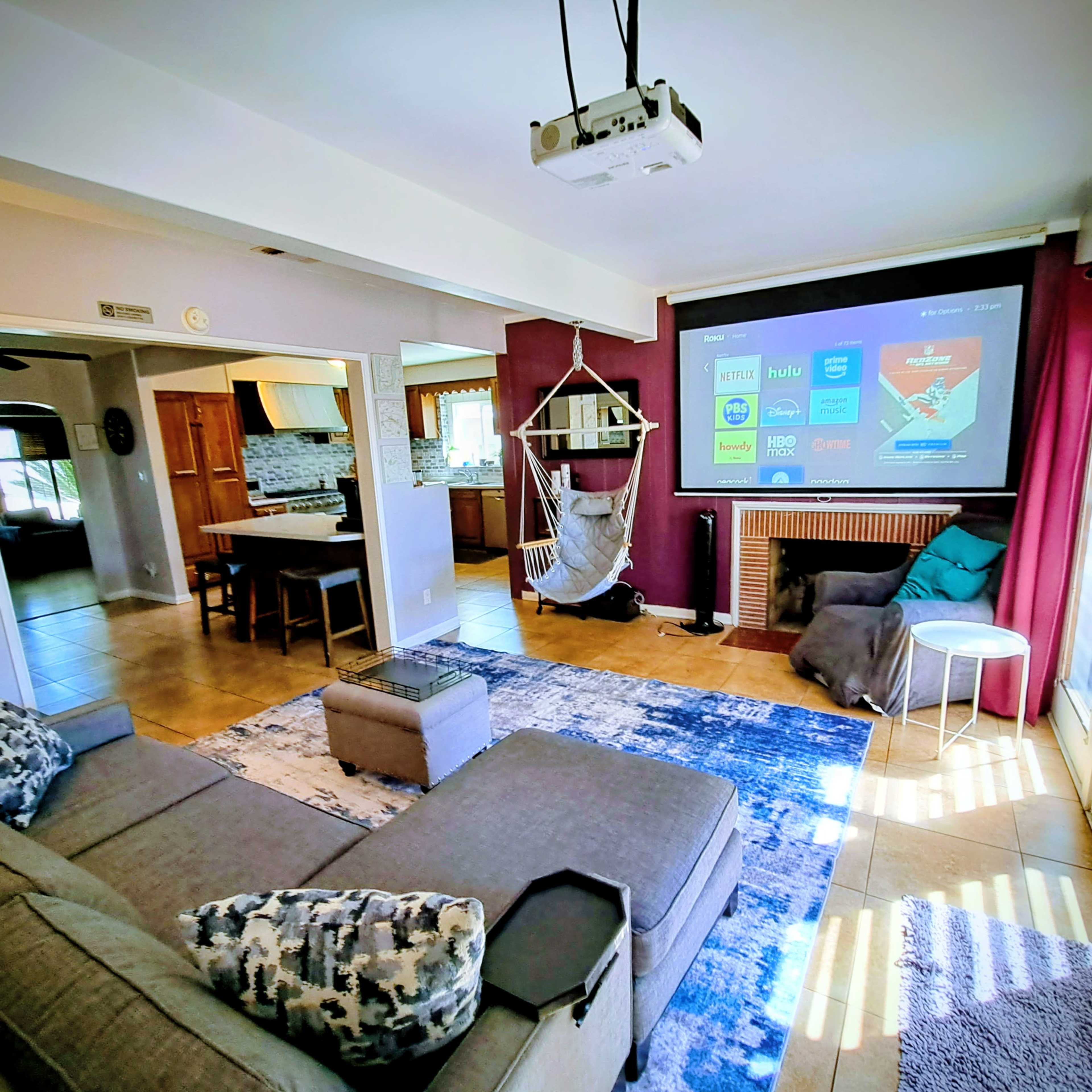 A modern living room features a projector screen displaying various streaming services, with a gray sofa, a swing chair, and a patterned rug on the tiled floor.