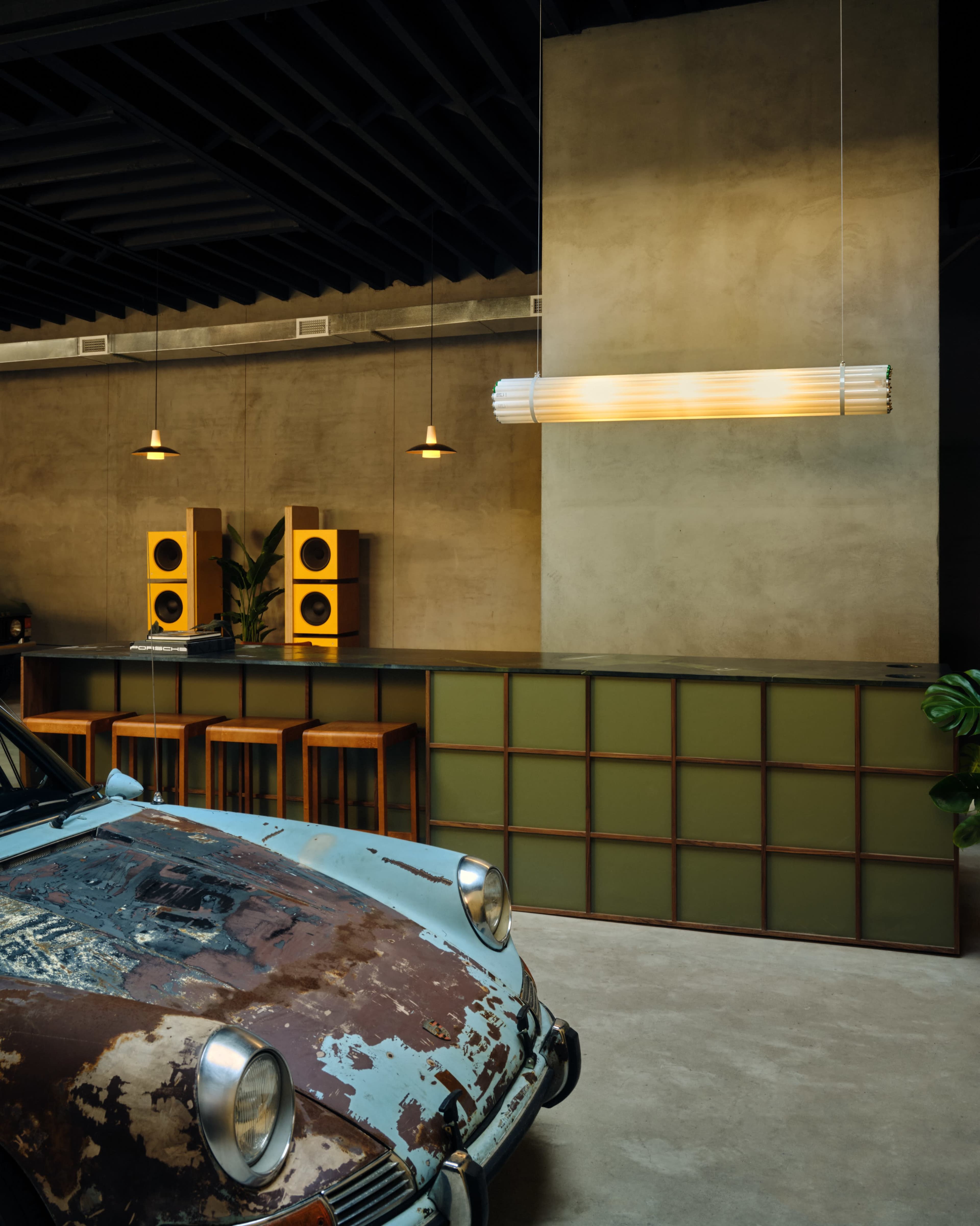 A vintage car with a weathered exterior is positioned in a modern interior space featuring a bar with stools, speakers, and pendant lighting.