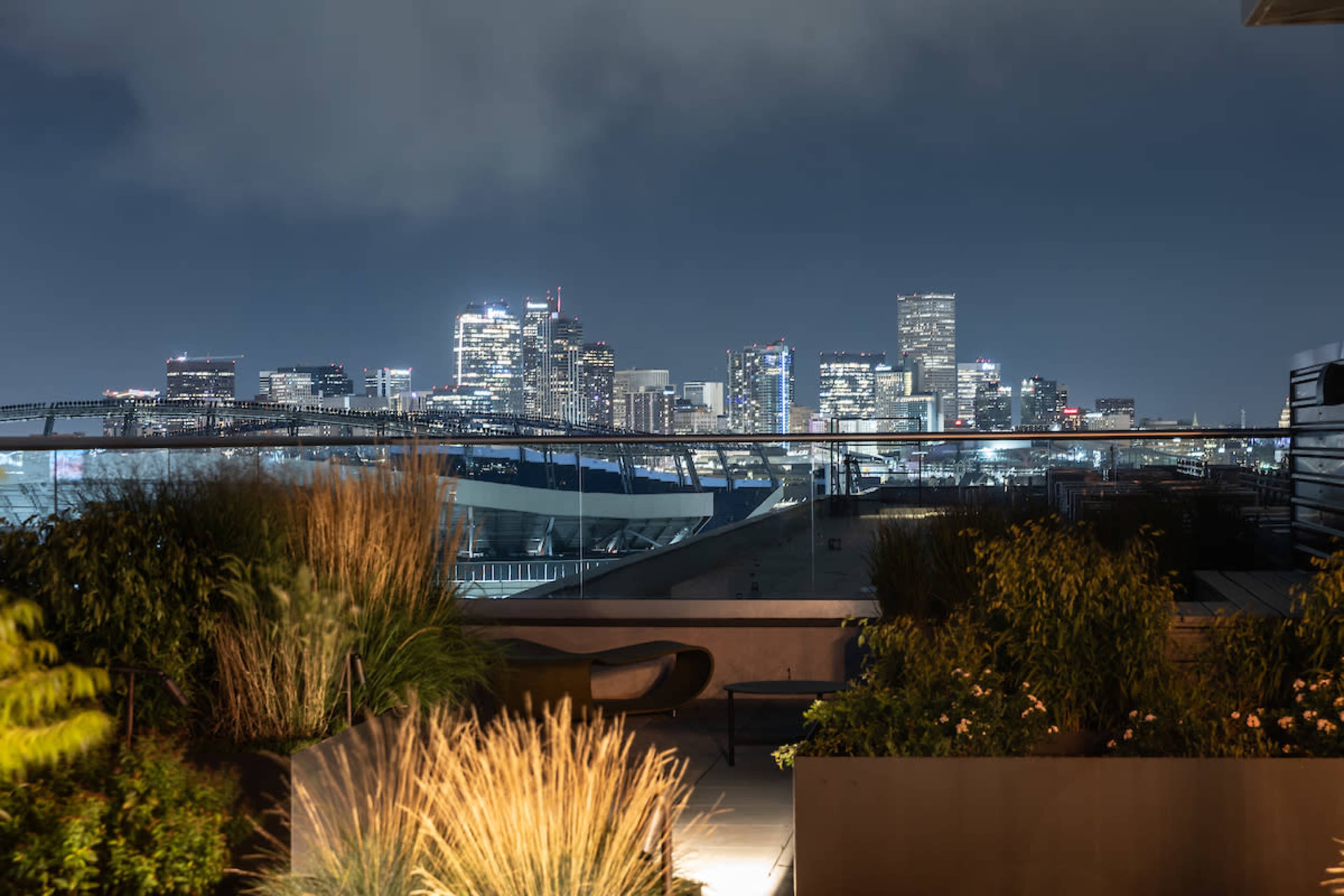 A rooftop garden overlooks a city skyline at night, with illuminated buildings and a sports stadium in the background.