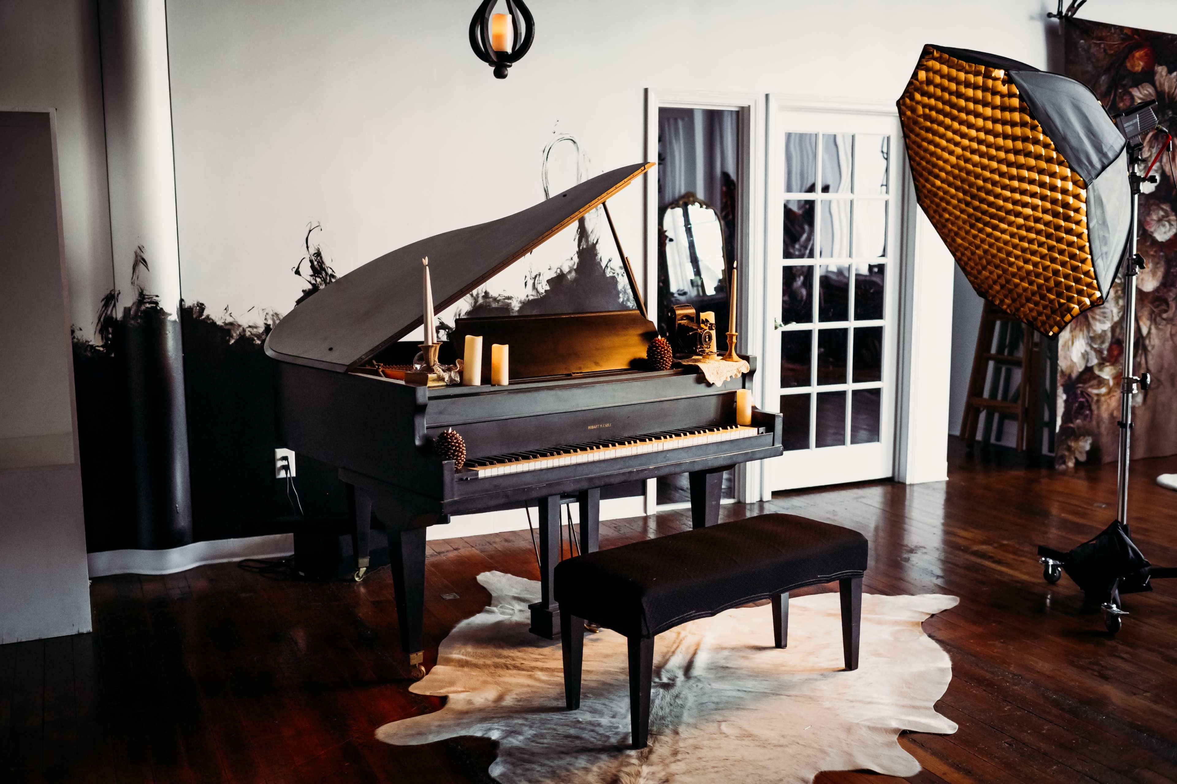 A black upright piano with a matching bench is positioned on a cowhide rug in a well-lit room featuring a large window and a photography light setup.