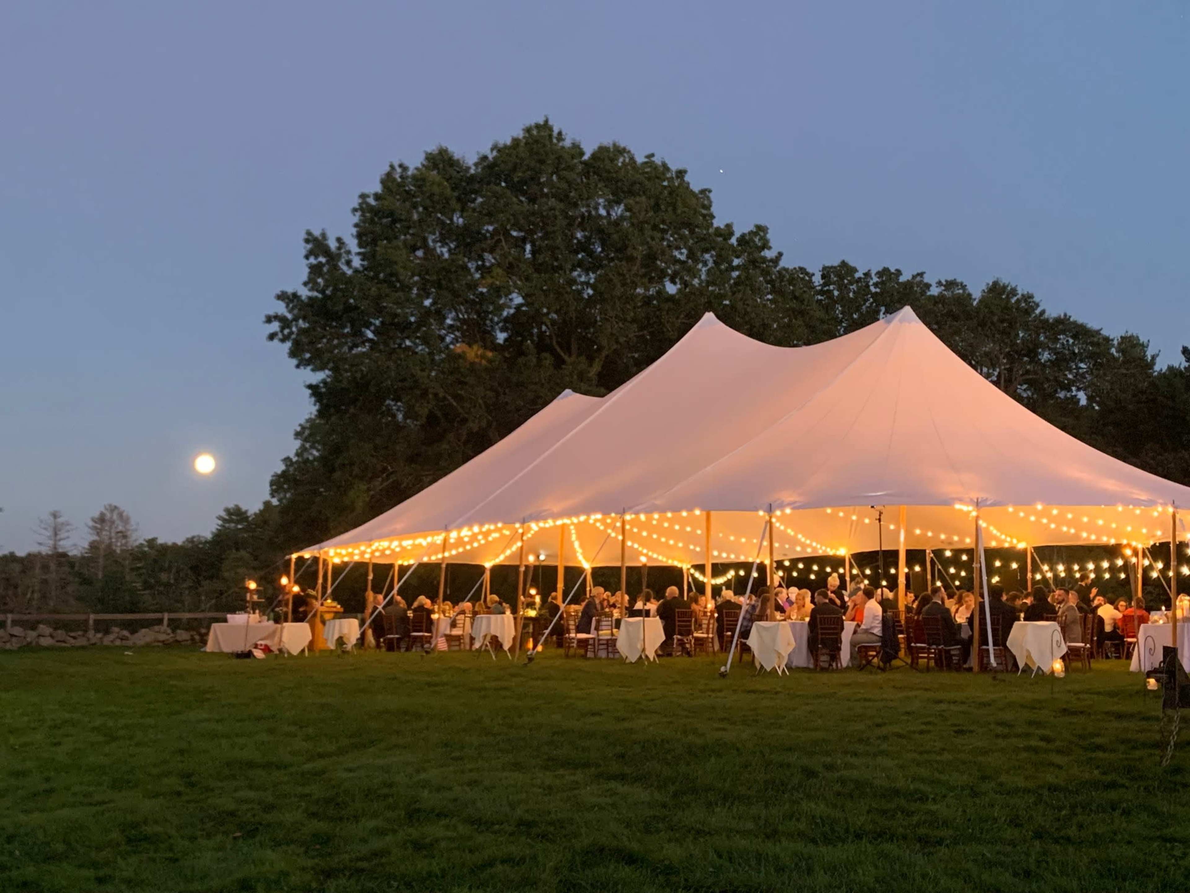 A large white tent decorated with string lights hosts an outdoor gathering on a grassy area near a tree line, with a bright moon visible in the sky.