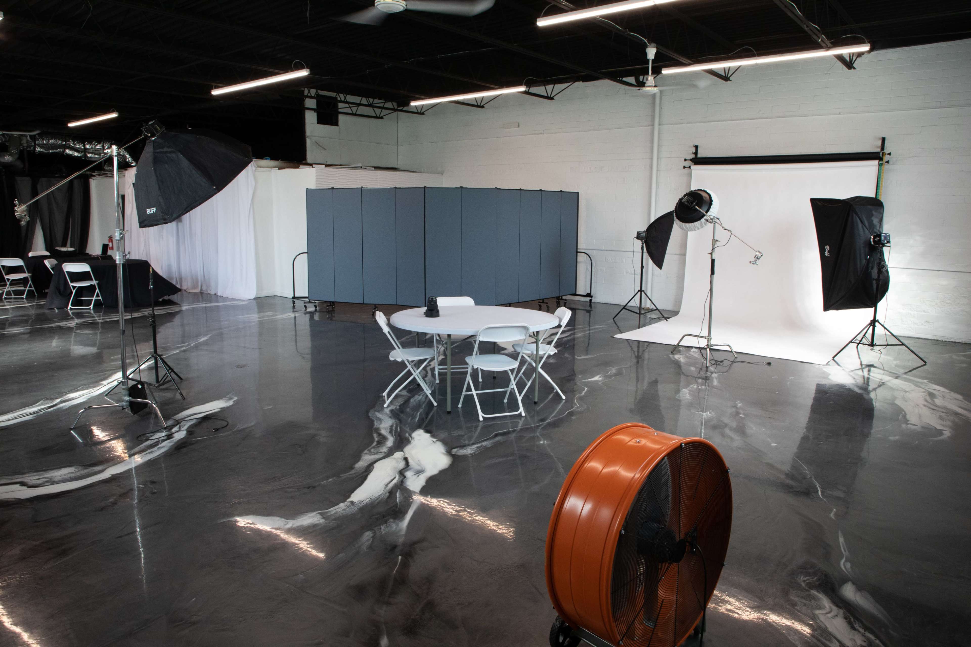 A spacious studio setup with a round table surrounded by chairs, photography equipment, and a large fan in a polished concrete environment.