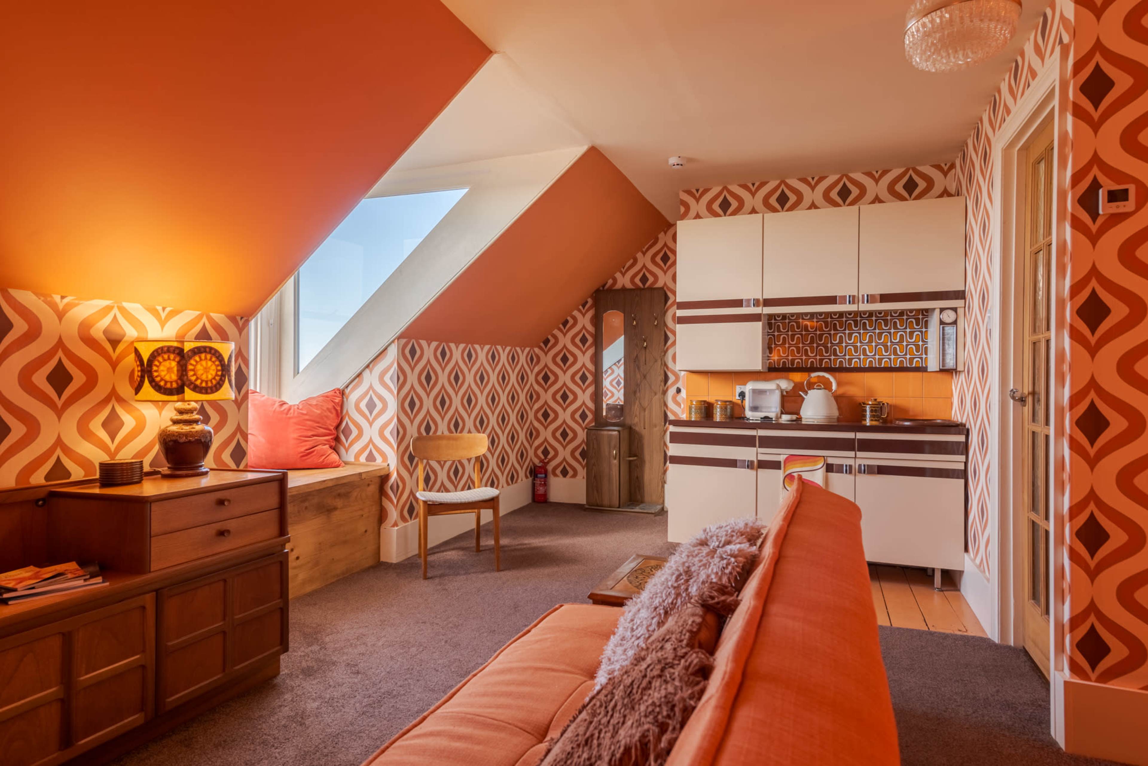 A penthouse room features orange geometric wallpaper, a kitchen area with retro cabinets, and a cozy seating arrangement.