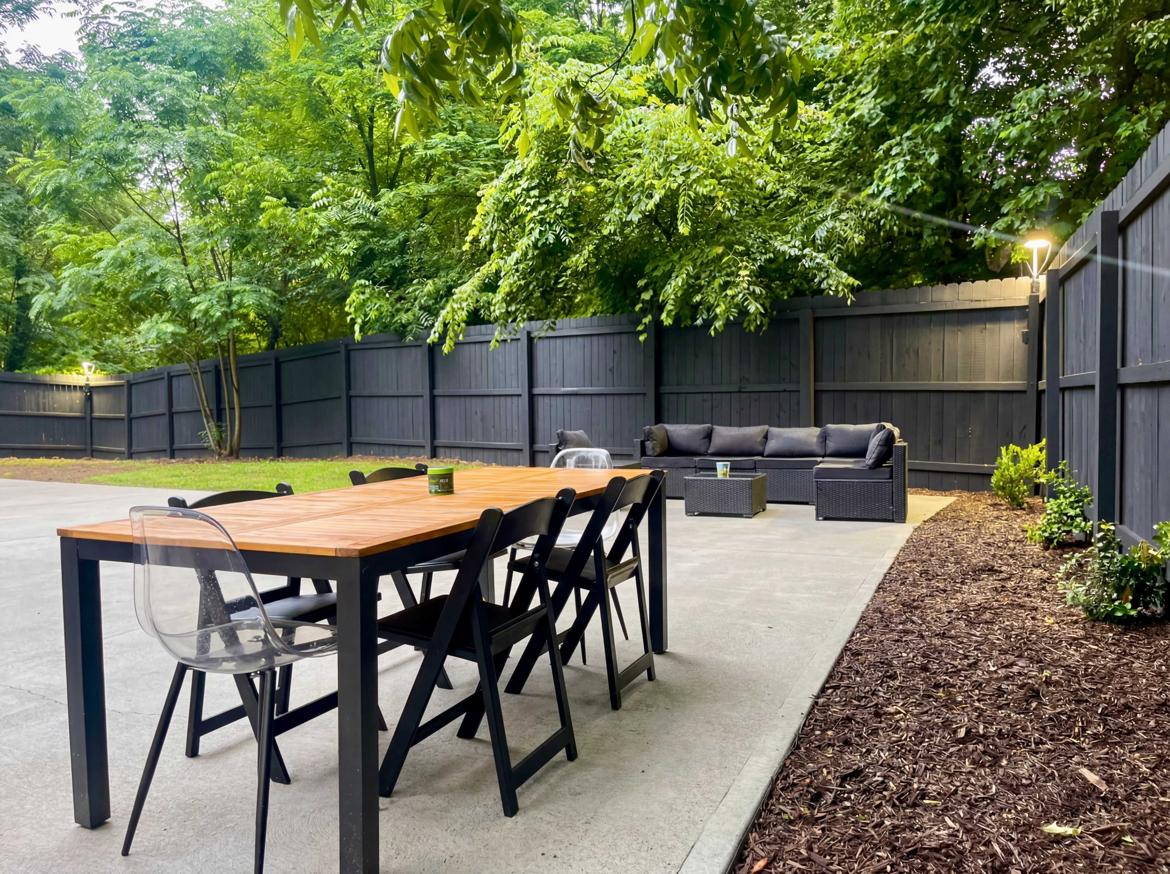The image shows a backyard patio featuring a wooden dining table with chairs, surrounded by green trees and a lounge area with a sofa.