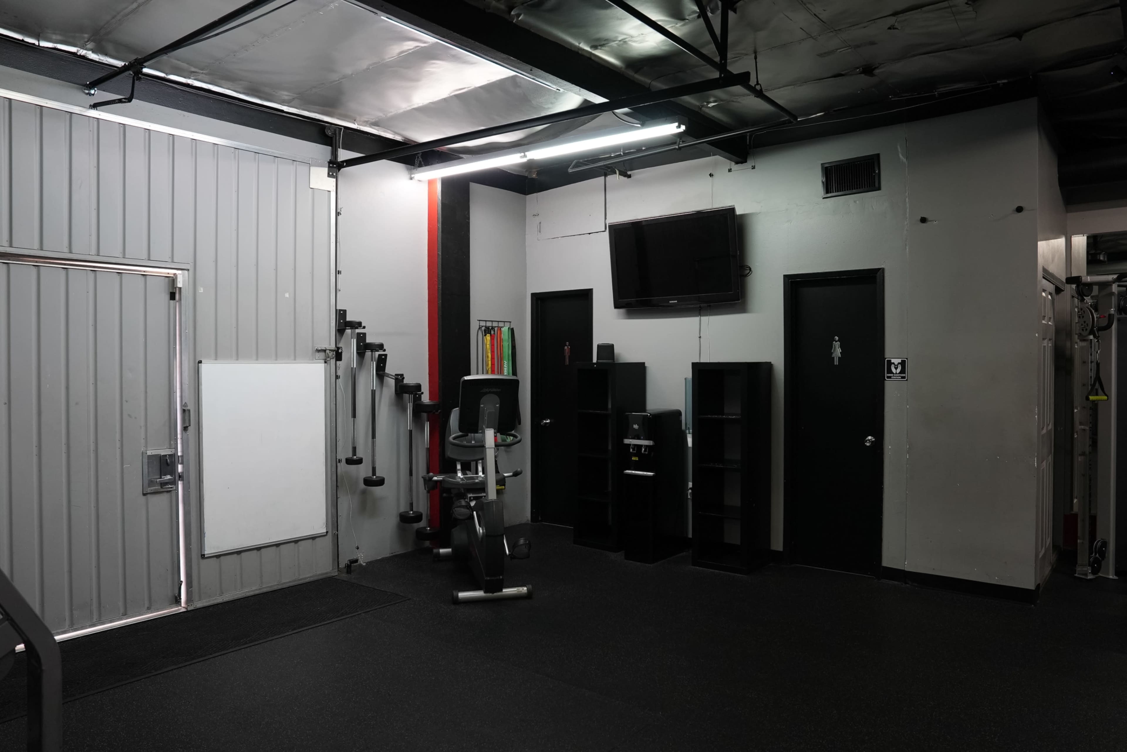 A corner of a gym featuring exercise equipment, a wall-mounted television, and storage shelves against a gray wall with a black door.