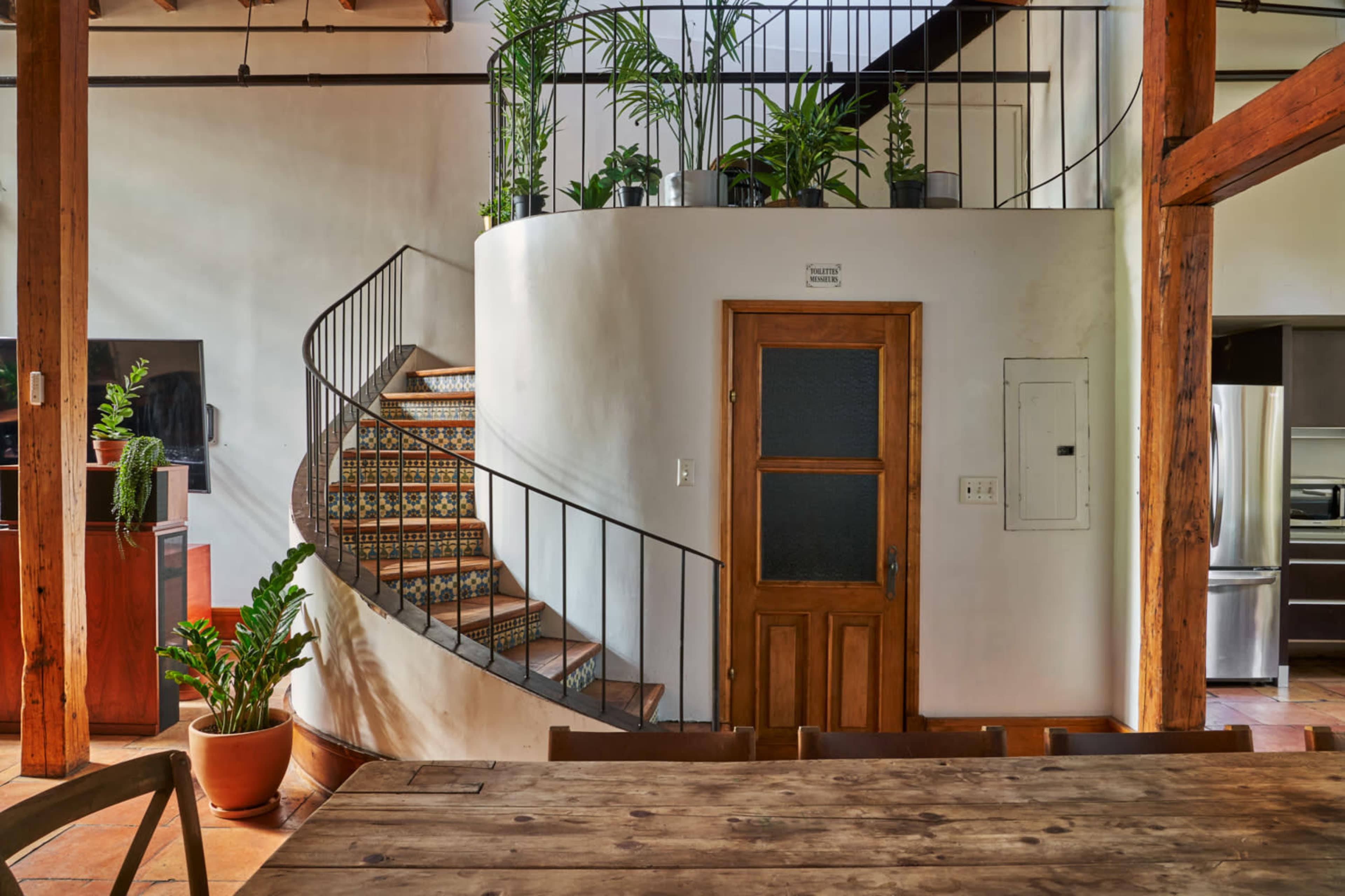 A spiral staircase with decorative tiles, flanked by potted plants, leading to a second level in a spacious interior with wooden beams and a dining table.