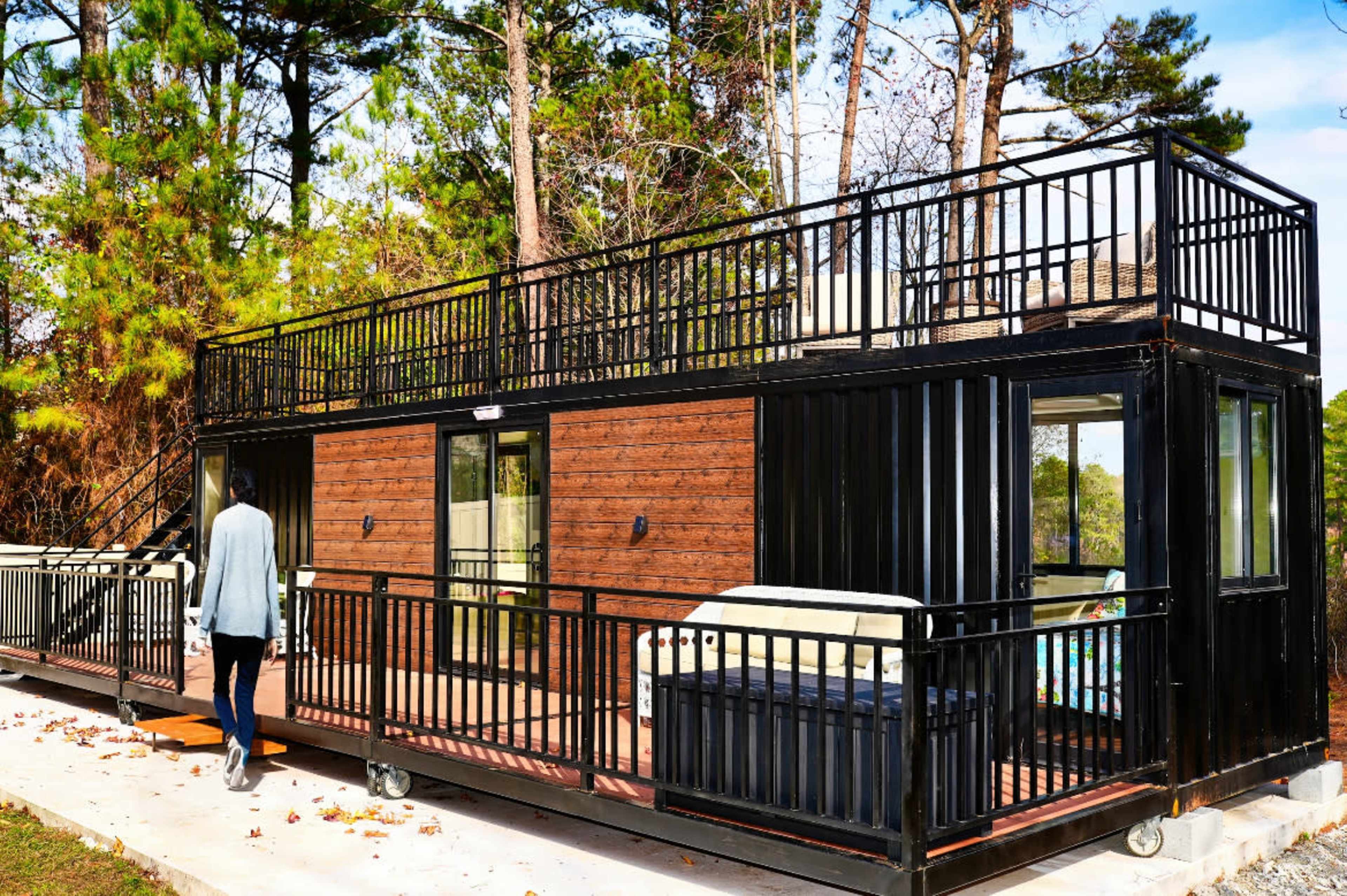 A person walks towards a modern, two-level container home with a rooftop deck situated among trees.