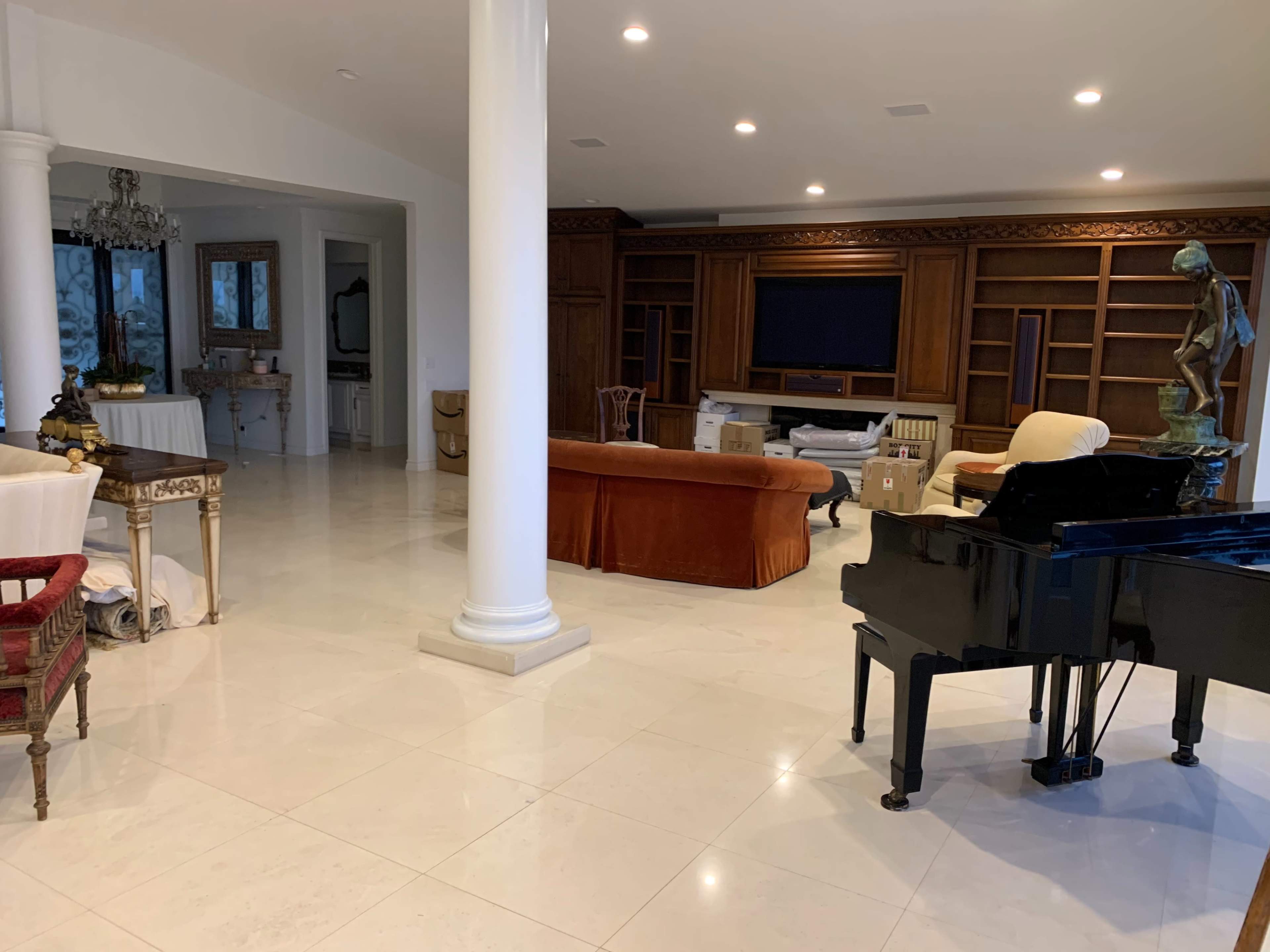 The spacious interior features a grand piano, a central column, and an arrangement of furniture, including a sofa and chairs, with shelves and a television in the background.