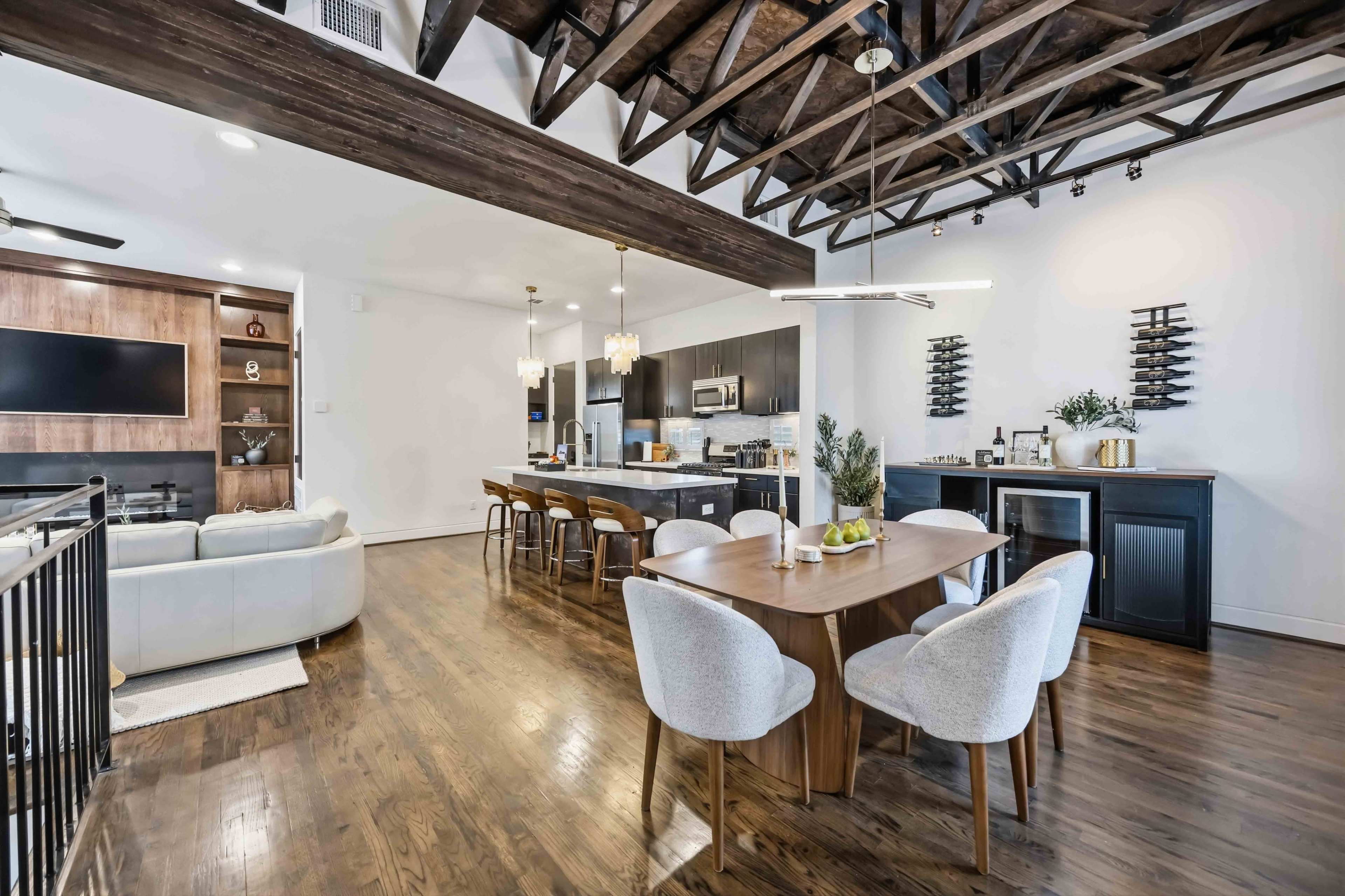 The image shows a modern open-concept living and dining area with a wooden dining table, sleek kitchen appliances, and exposed wooden beams on the ceiling.