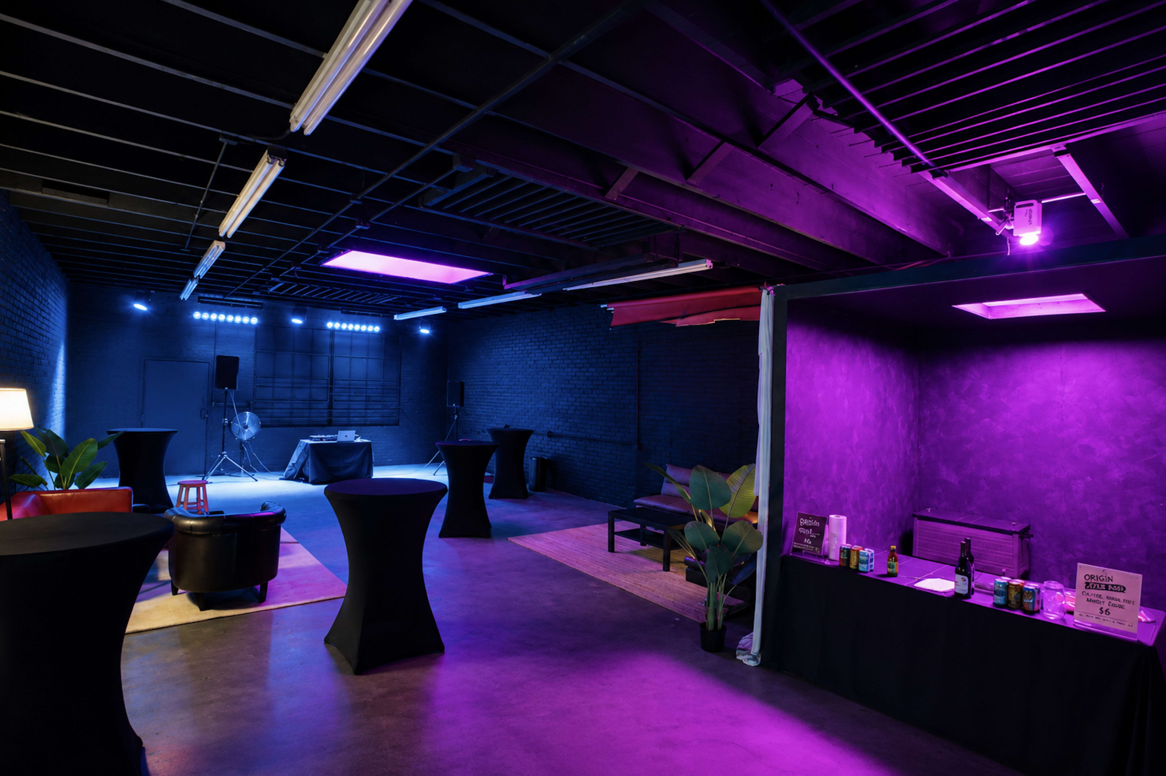 The image shows a dimly lit event space featuring purple and blue lighting, with tables arranged around a central area and a small bar setup along one wall.