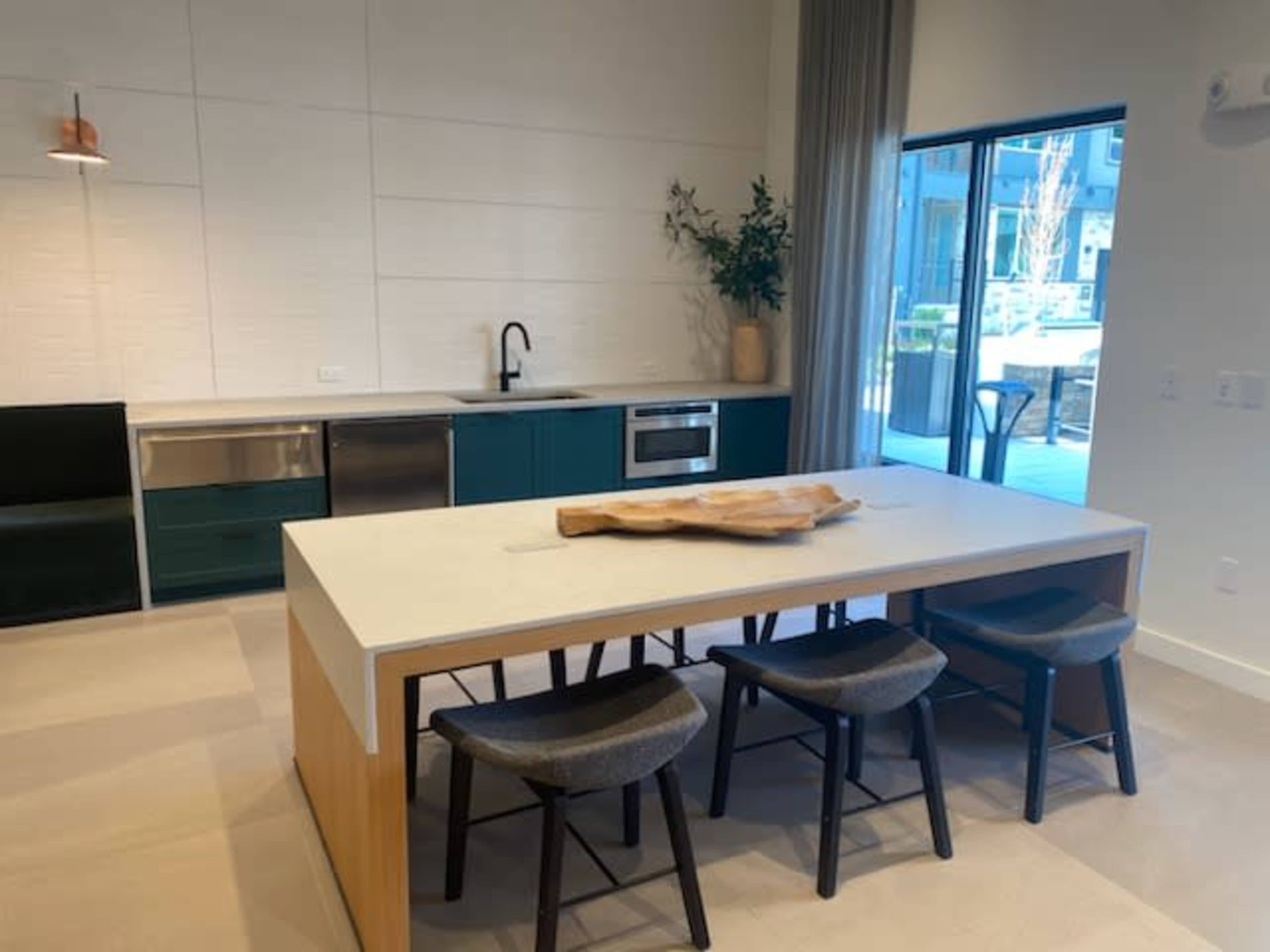A modern kitchen features a large wooden dining table with four black stools, a green and silver cabinetry, and large windows that allow natural light to enter.