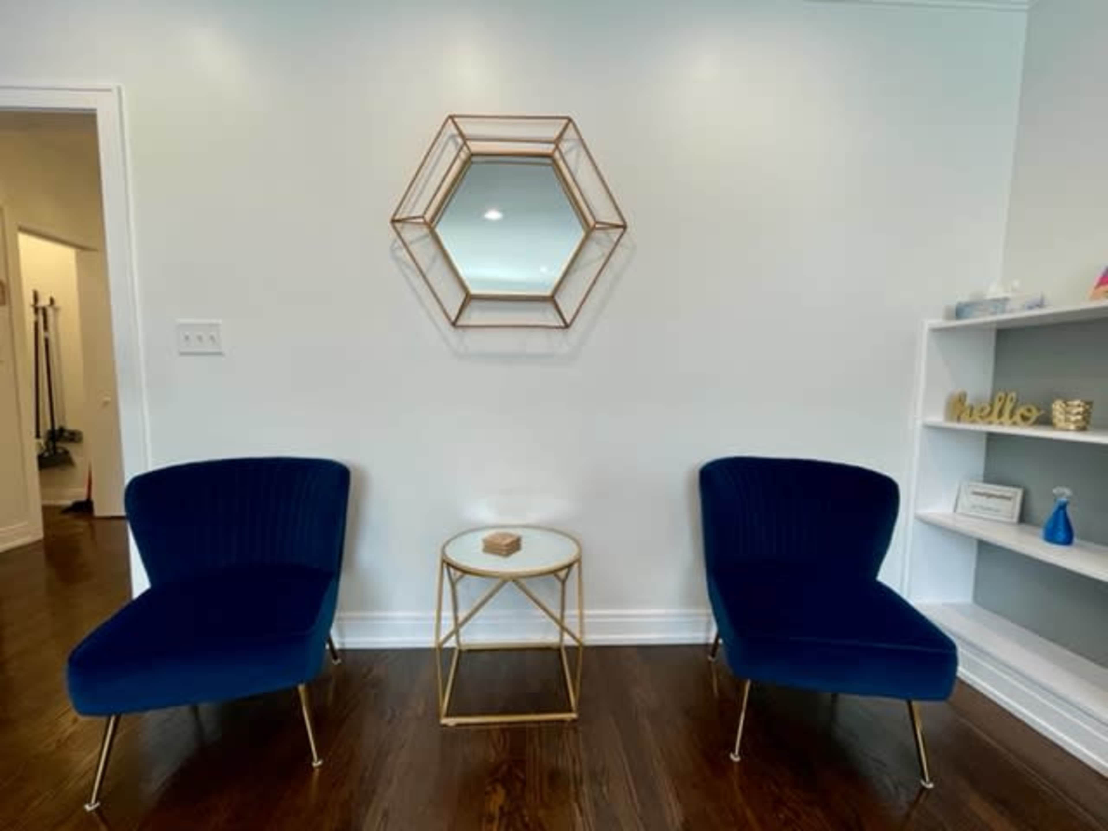 Two blue chairs are positioned facing a small gold side table and a hexagonal mirror on a white wall.