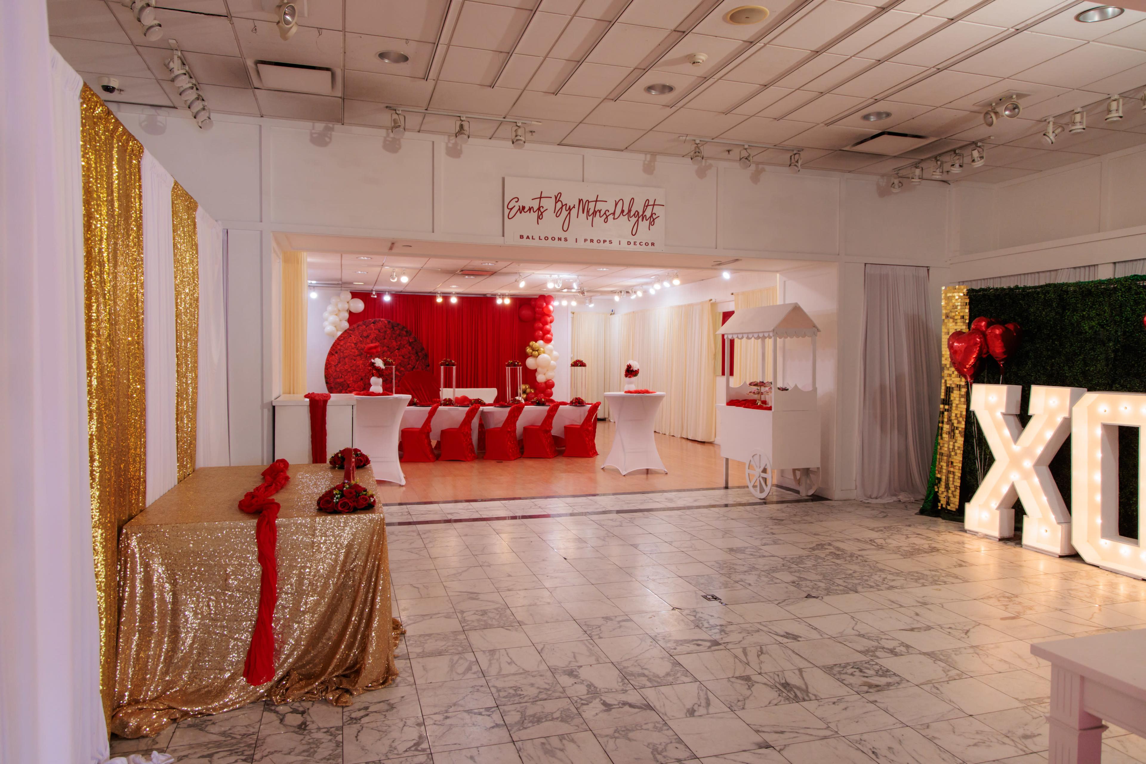 The image shows a decorated event space with gold and red accents, featuring a table with a glittery tablecloth, chairs arranged in rows, and large illuminated letters spelling "XO."