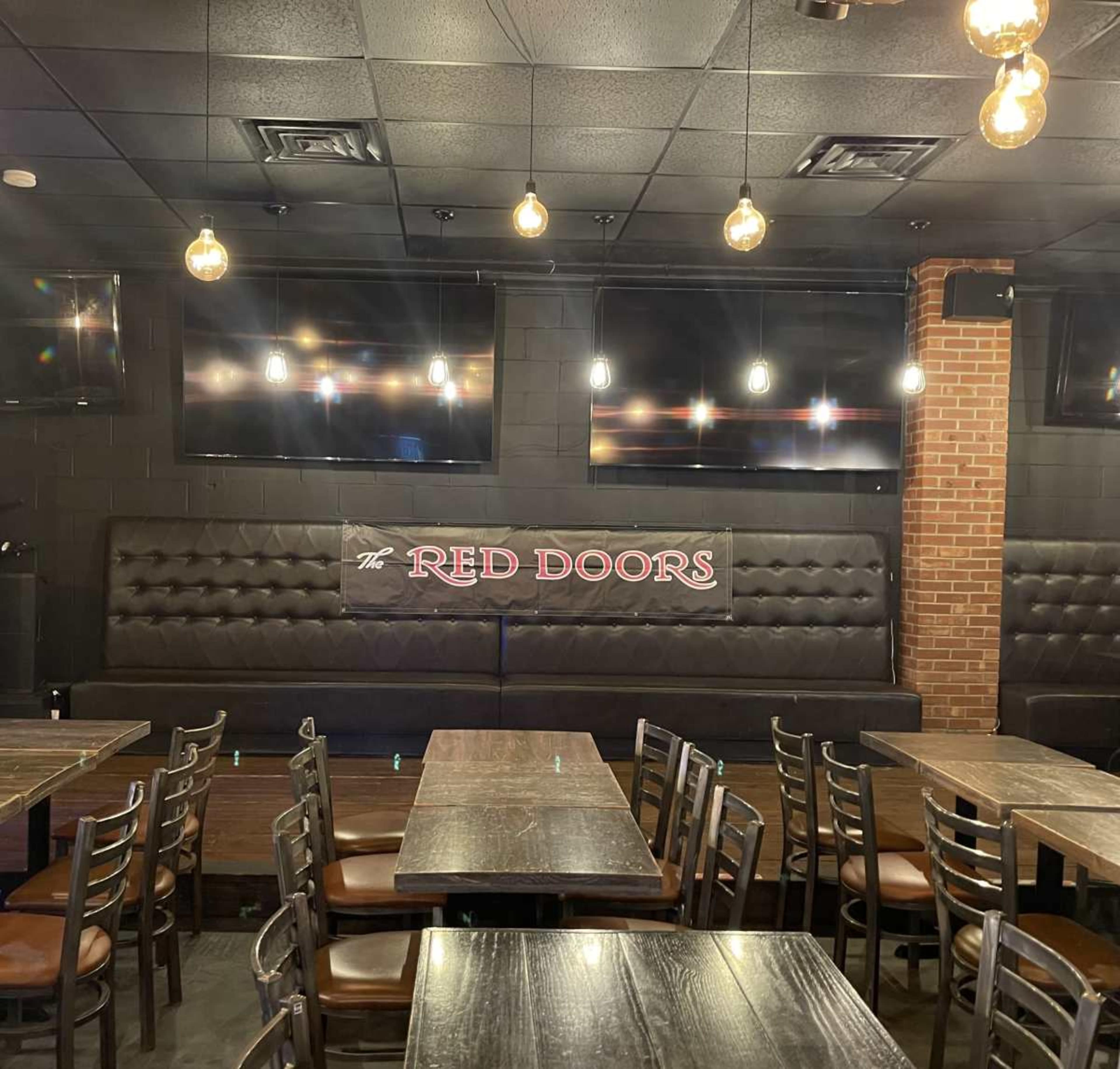 The image shows the interior of a restaurant with a black and red color scheme, featuring rows of wooden tables and chairs, a long padded seating area, and multiple flat-screen televisions mounted on the walls.