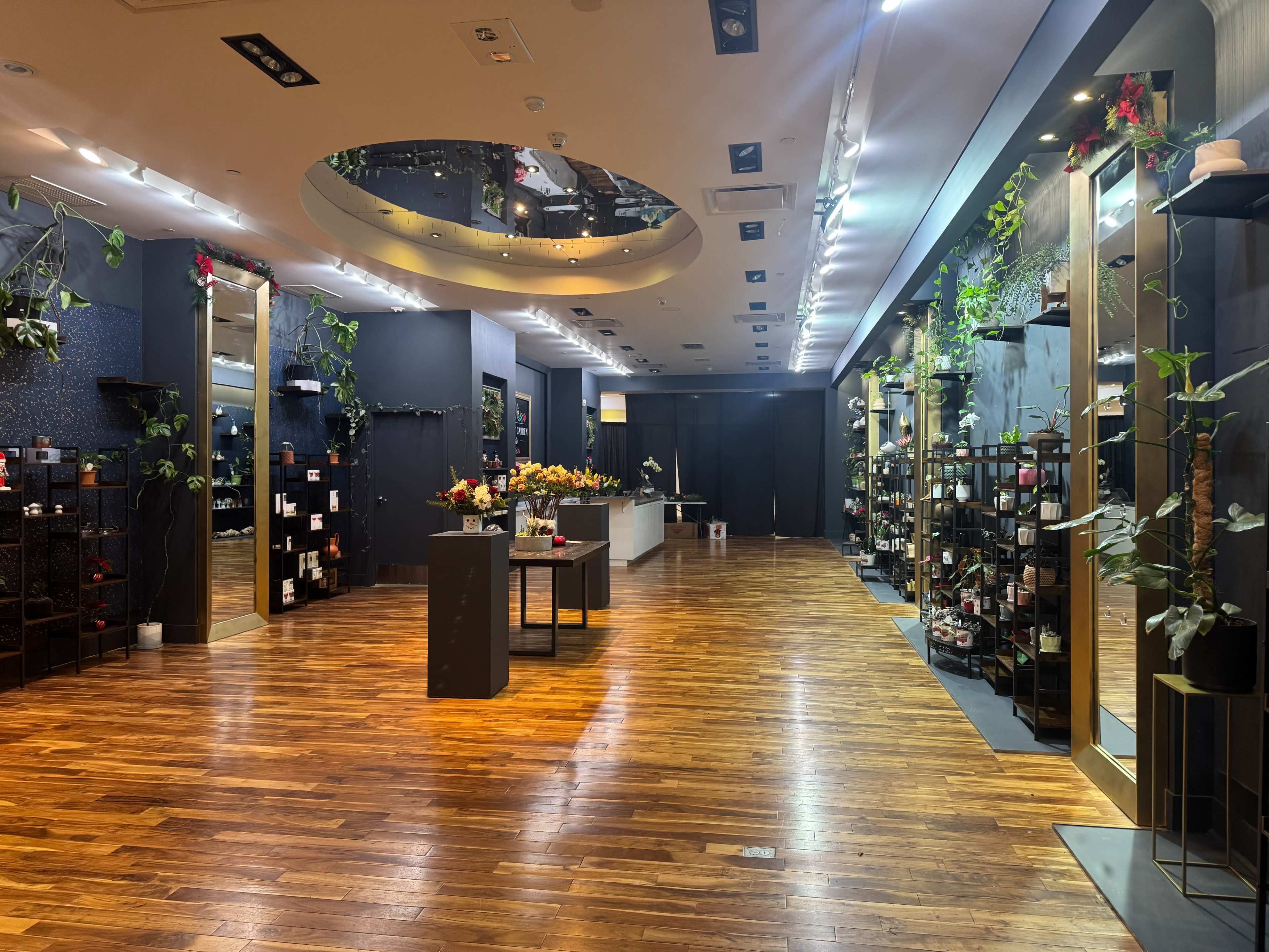 The image shows a spacious retail store with wooden flooring, illuminated displays, and shelves lined with various products, adorned with potted plants and floral arrangements.