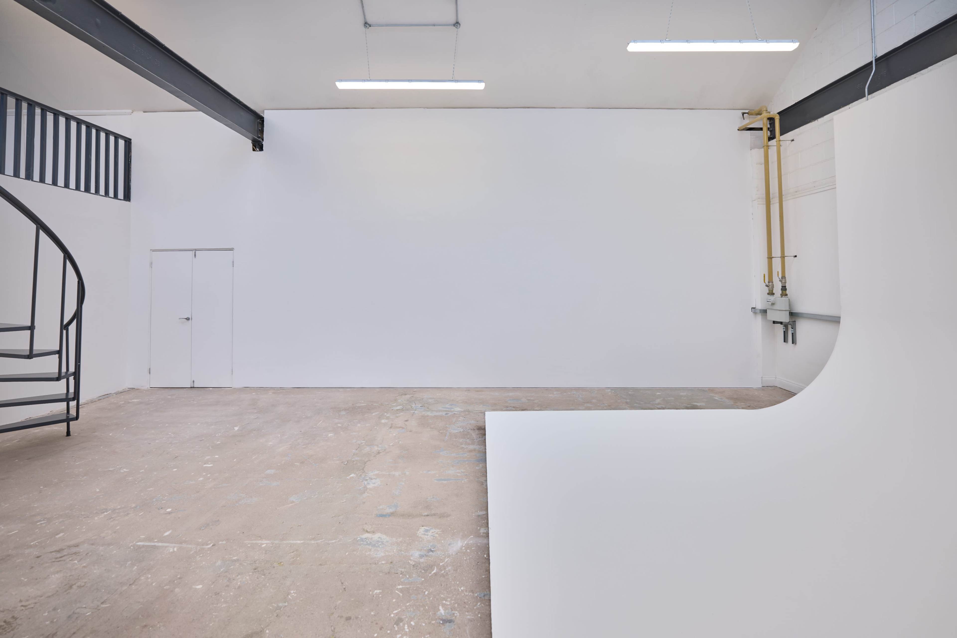 The image shows an empty, bare studio space with a curved white backdrop, concrete floor, and a spiral staircase in the background.