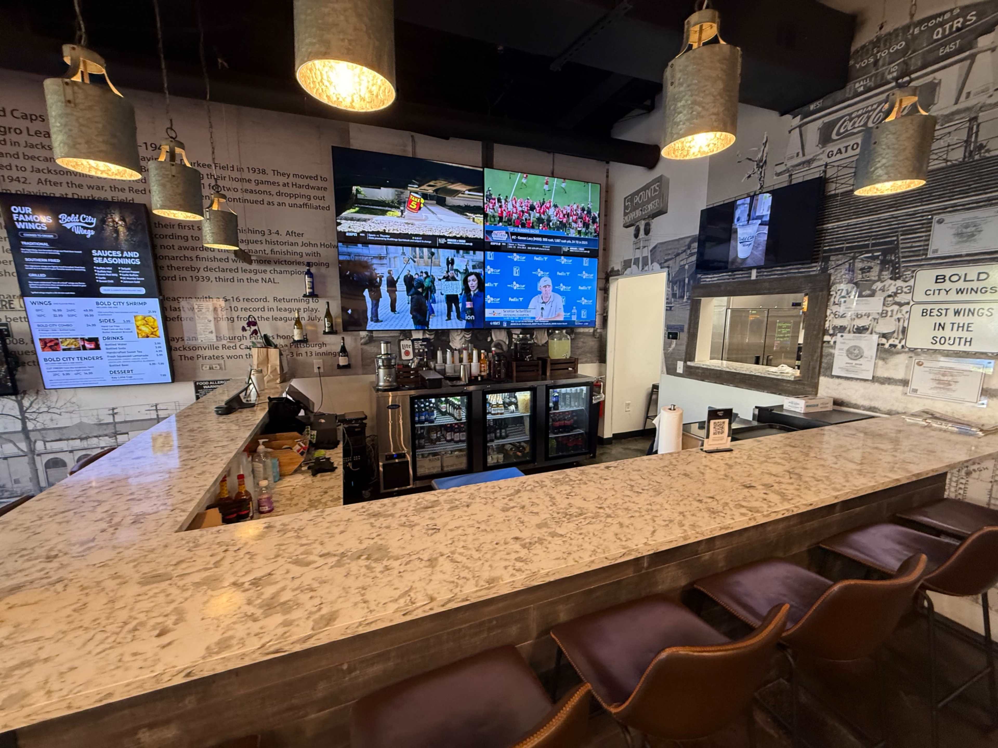 The image shows a bar area with a granite countertop, rows of brown chairs, multiple screens displaying sports, and a refrigerator filled with drinks.