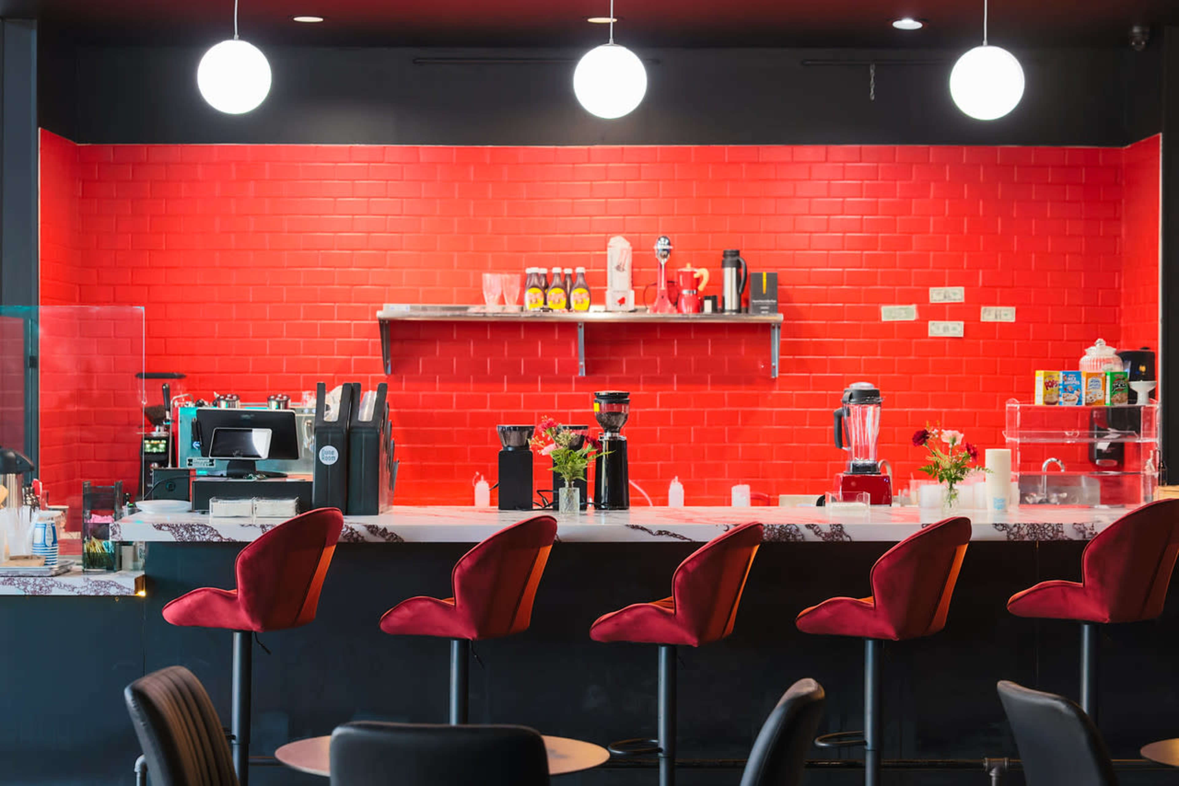 A modern cafe features a bright red wall, a countertop with bar stools, and various appliances displayed on shelves.