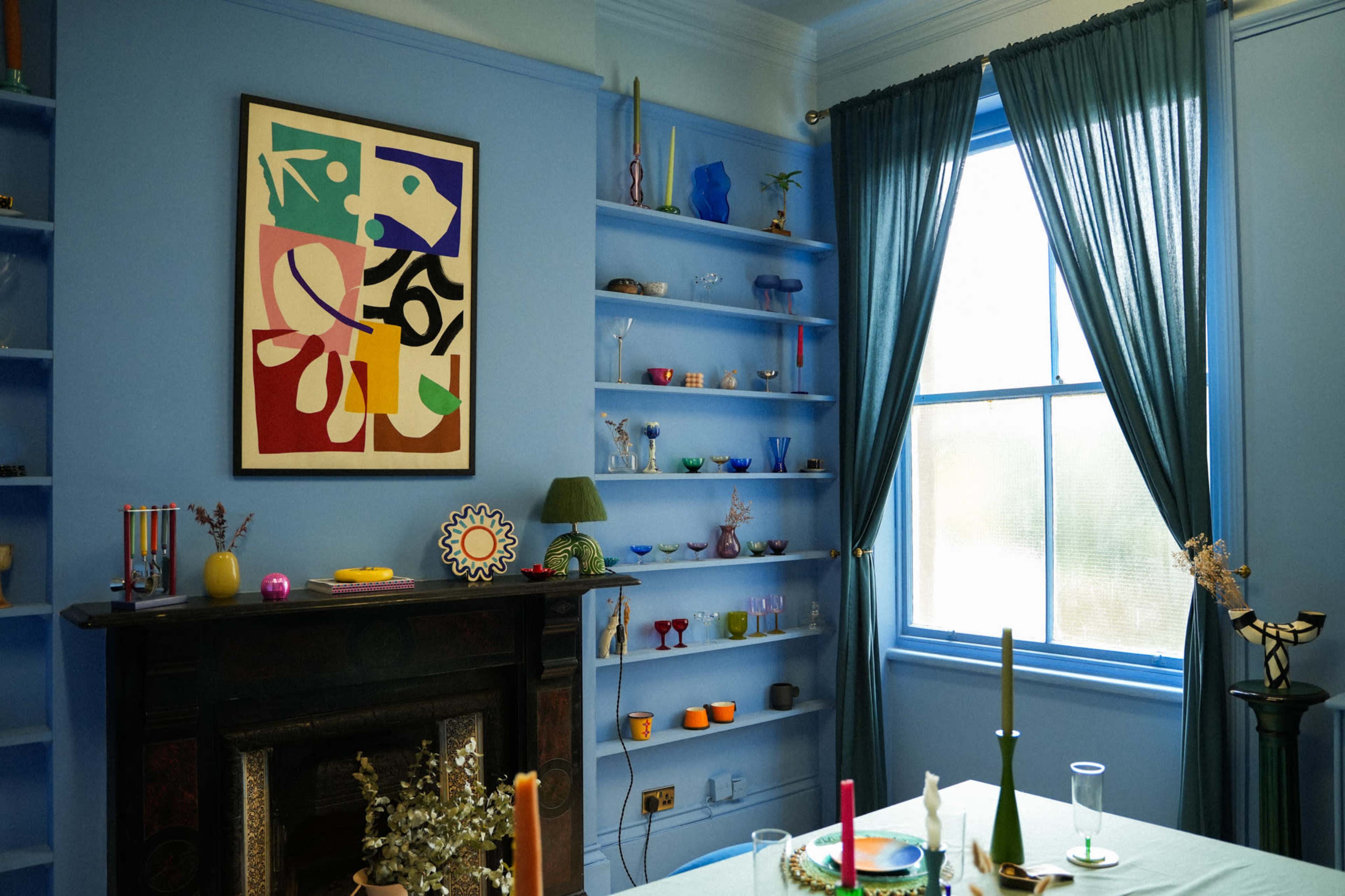 The room features a blue-painted wall adorned with colorful artwork and shelves displaying various decorative items, alongside a table set with candles and a vase of flowers.